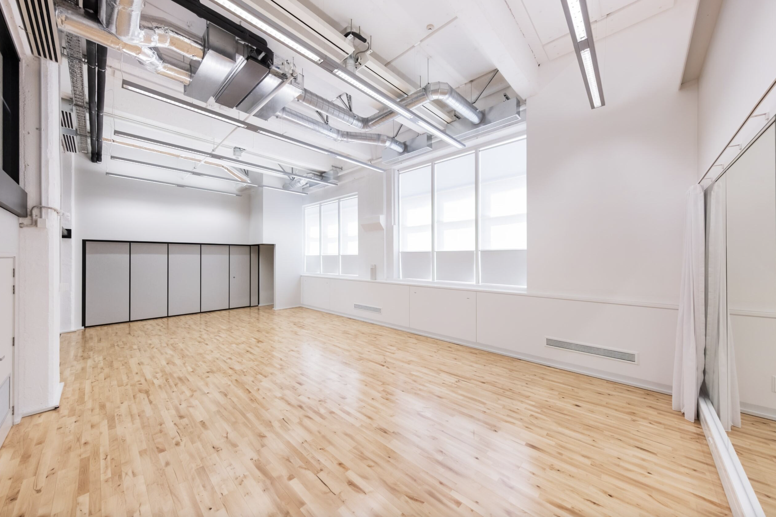 Empty room with wooden flooring, white walls, large windows, and exposed silver ductwork on the ceiling.