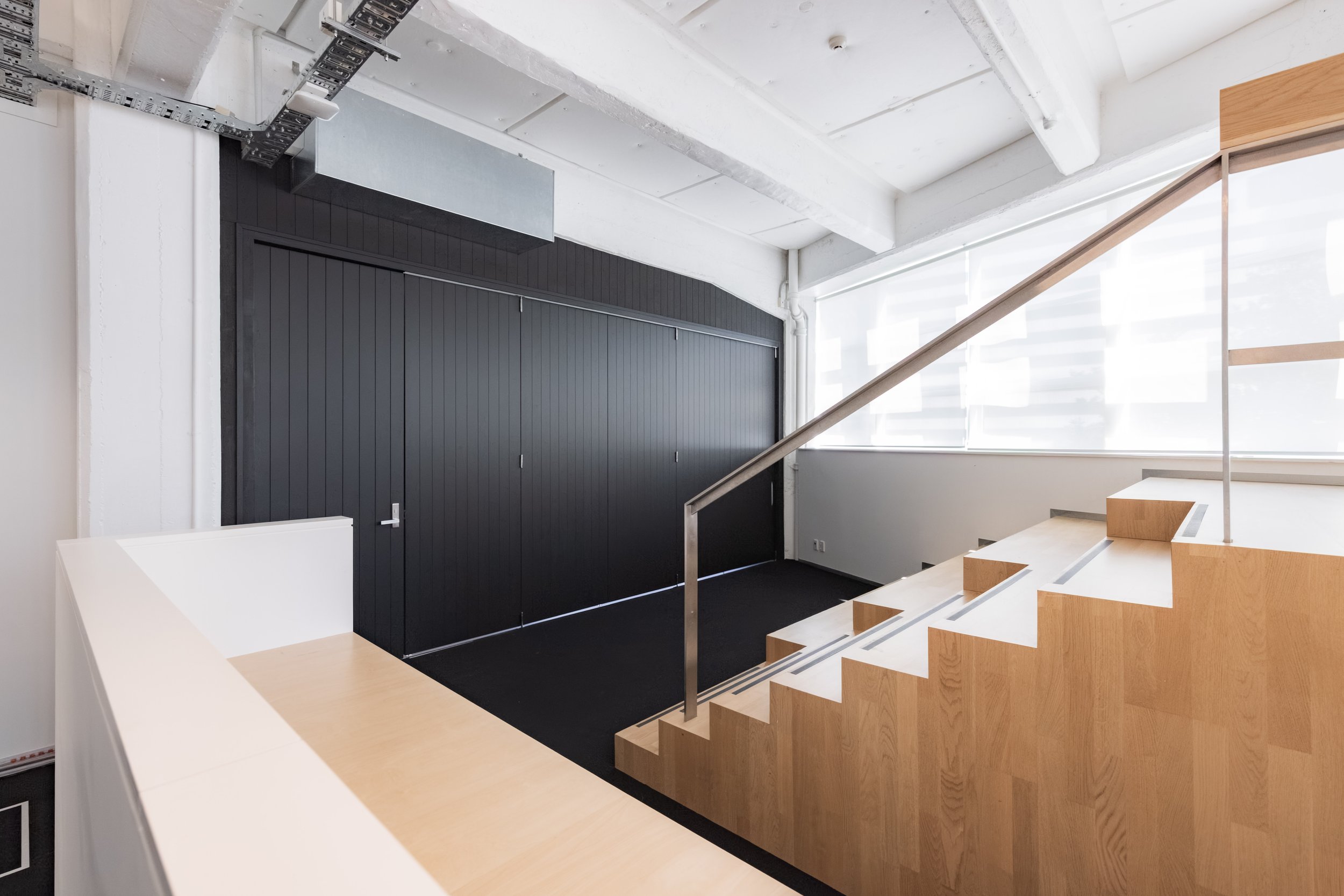 Toi Aro: Interior view of a modern staircase with light wooden steps and a metal railing, next to a black sliding door, with a white ceiling and walls decorated with exposed pipes and industrial elements.