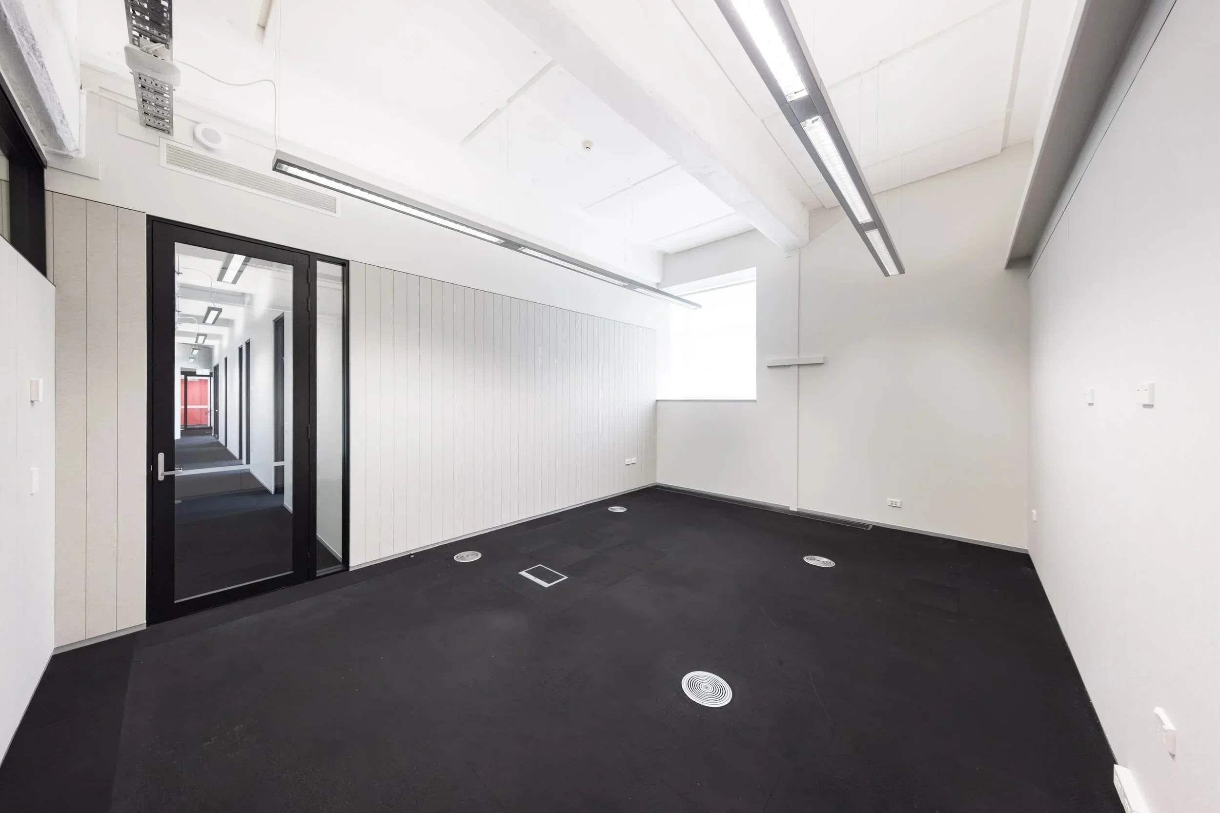 Te Hau Āwhiowhio - an empty office space at Toi Aro. 30 m² with a capacity of up to 6 people. White walls, dark grey carpet, office with partition walls, glass windows and door.