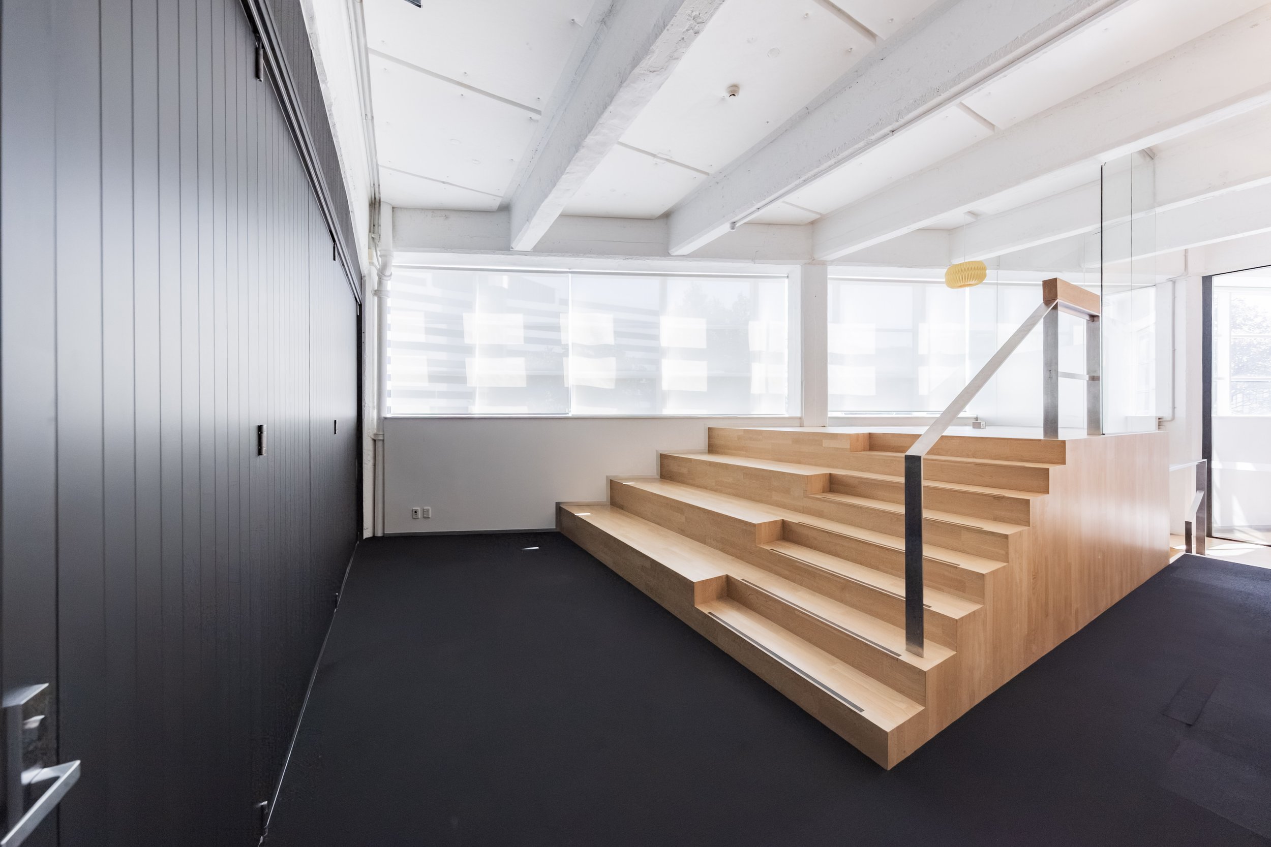 Toi Aro: Interior view of a modern space with white ceiling and walls, large windows with shades, and a wooden staircase with a black railing.