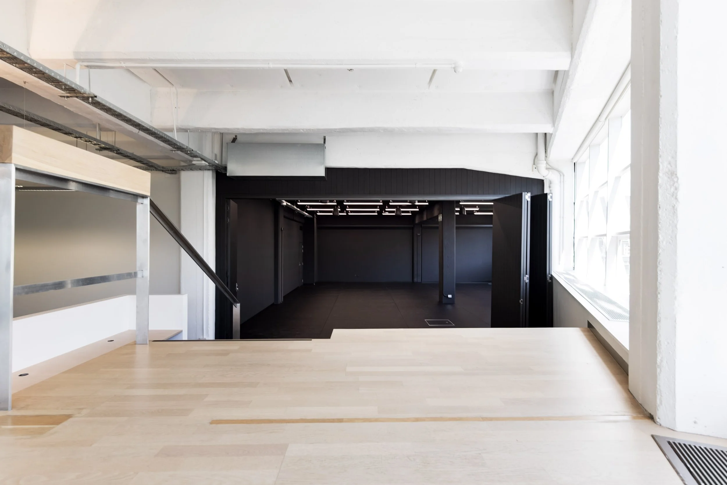 Toi AroInterior view of a modern commercial space with light wooden flooring, large windows on the right, and a dark-colored room or corridor at the back. The ceiling features exposed beams and ductwork, with natural light coming through the windows.