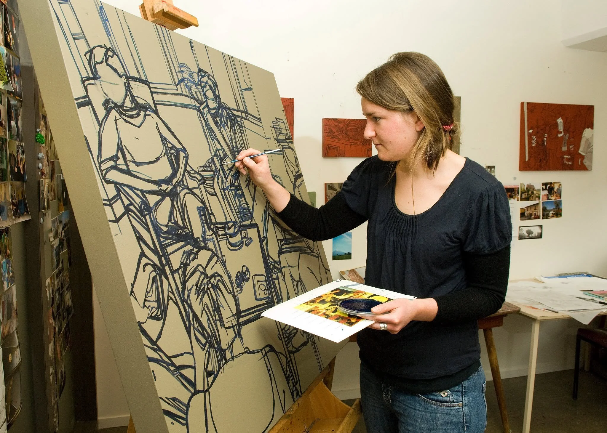 Female painting in black paint on brown cardboard on an easel.