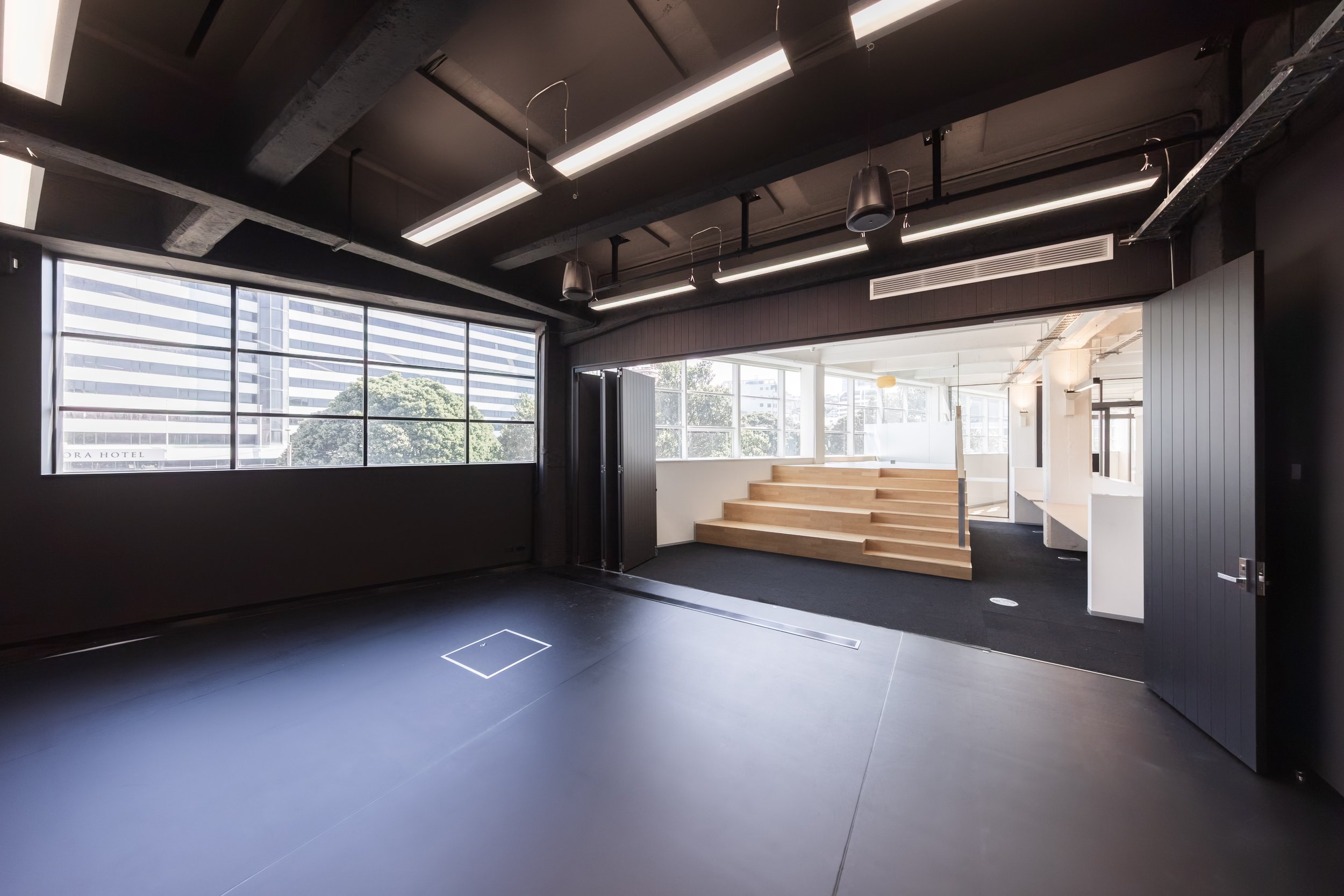 Toi Aro: An interior view of a modern, minimalist office or conference room with large windows letting in natural light, black walls and ceiling, illuminated ceiling fixtures, and a wooden staircase leading to a raised platform.