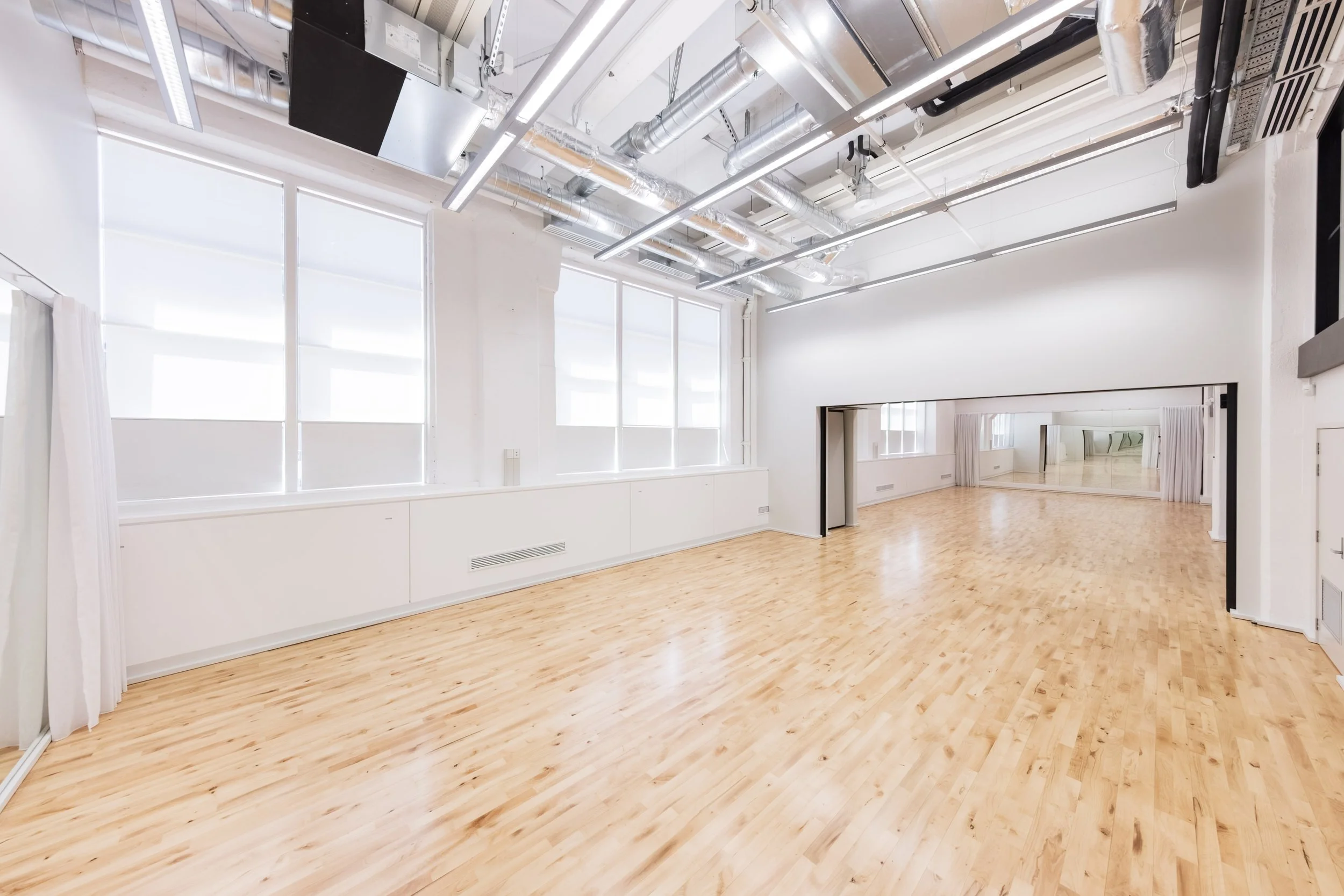 Empty dance or fitness studio with large windows, wooden floor, and ceiling industrial ductwork.