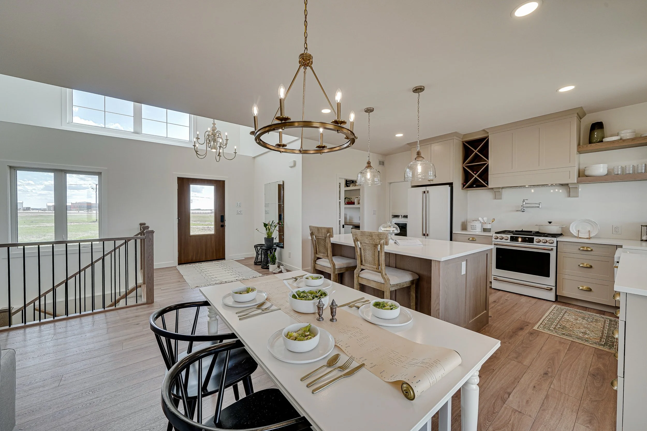 A modern, open-concept kitchen and dining area with a white table set with bowls of salad, black chairs, and a kitchen with beige cabinets, a white stove, and open shelves displaying dishes. Large windows provide natural light, and there are decorative light fixtures hanging from the ceiling.