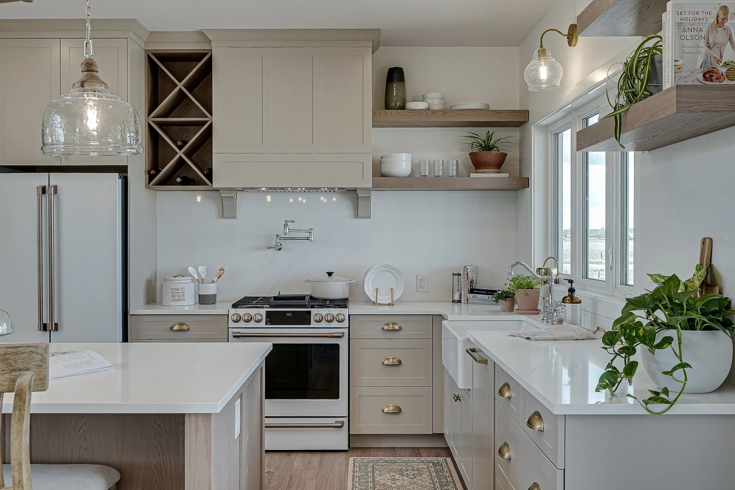 A modern kitchen with off-white cabinets, a stainless steel stove, a white farmhouse sink, and open wooden shelves with plants and dishes. There is a window above the sink, and a glass pendant light hangs above. The countertop has a potted plant, soap dispenser, and a dish towel. The kitchen island has a book and some dishes on it.