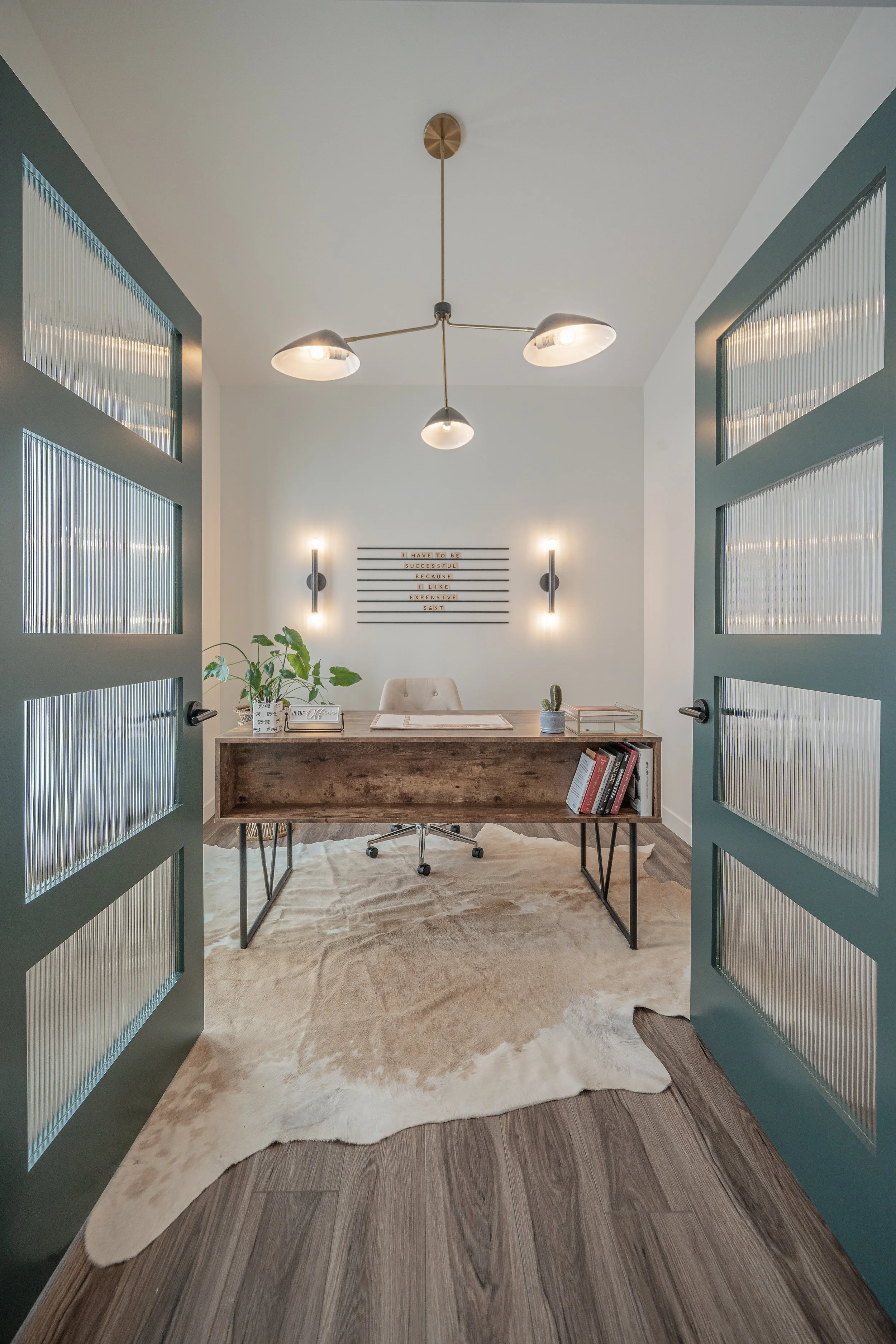 Home office with a wooden desk, chair, plants, bookshelf, and wall décor, seen through an open door with glass panels, on a wooden floor with a cowhide rug.