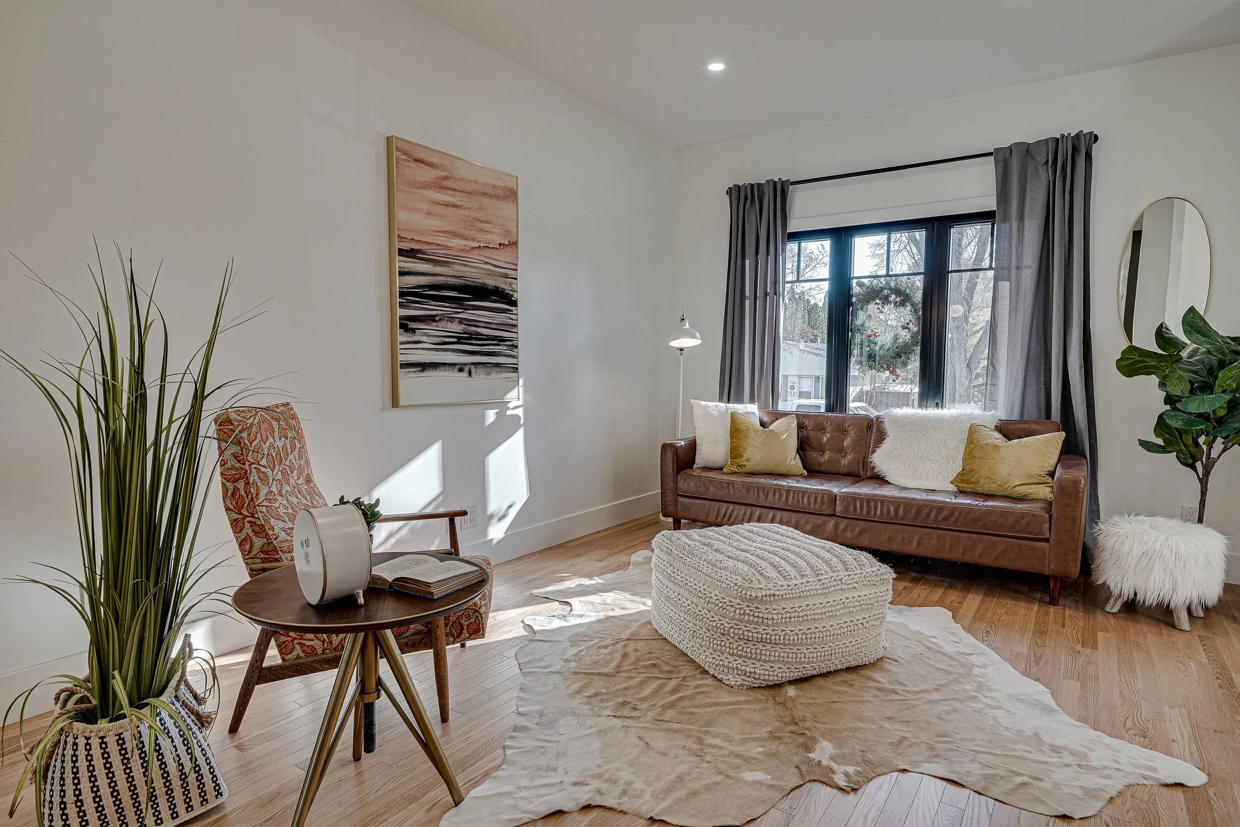 A cozy living room with a leather sofa, throw pillows, a pouf, a cowhide rug, a small round wooden table with an open book, a potted plant, a wall painting with abstract pink and black stripes, a tall floor lamp, a large window with dark curtains, and a mirror on the wall.