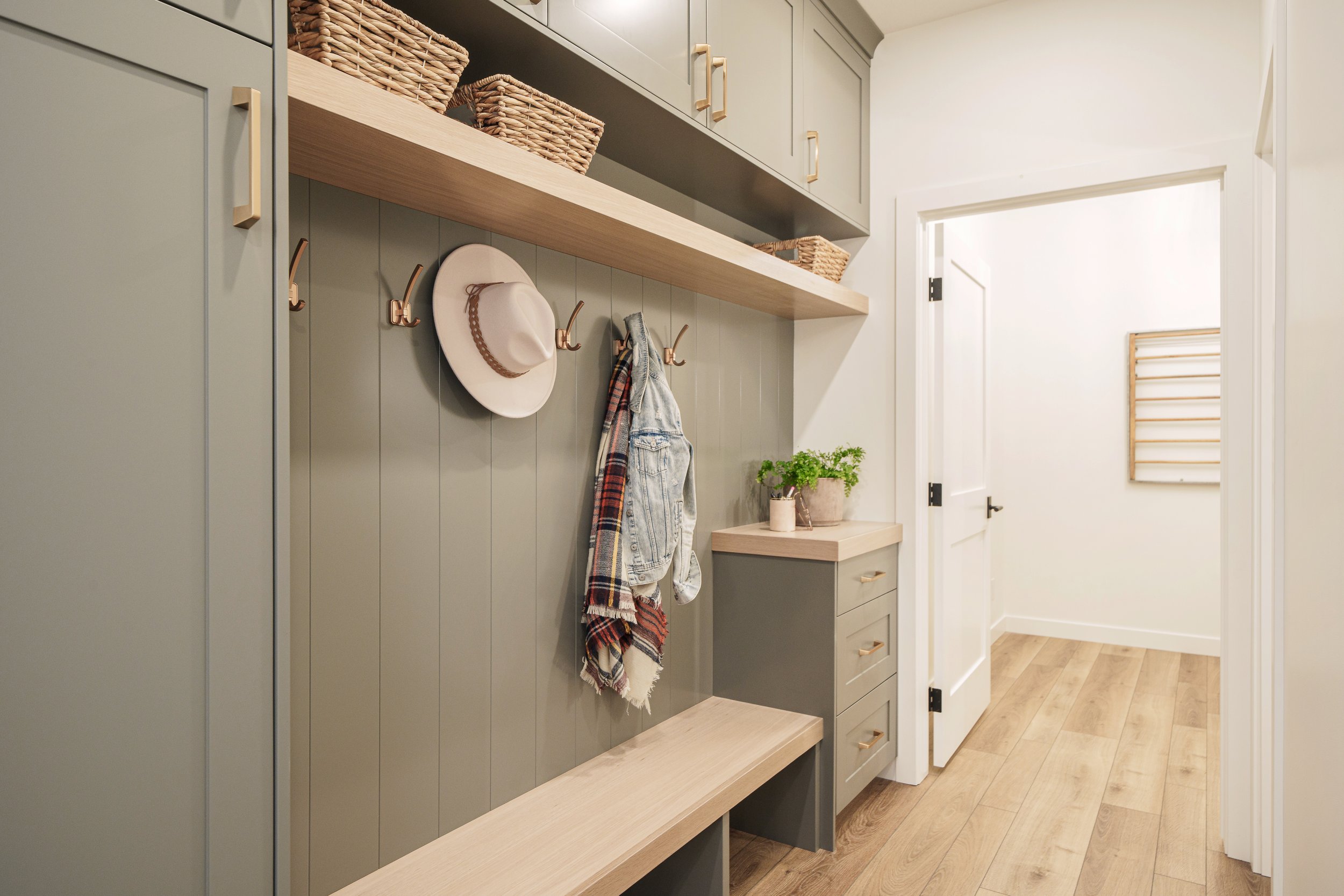 A hallway with built-in storage units, including cabinets, hooks with hats and scarves, and a shelf with baskets, along with a small table holding a potted plant. The floor is hardwood, and the walls are painted in a light gray and white color.