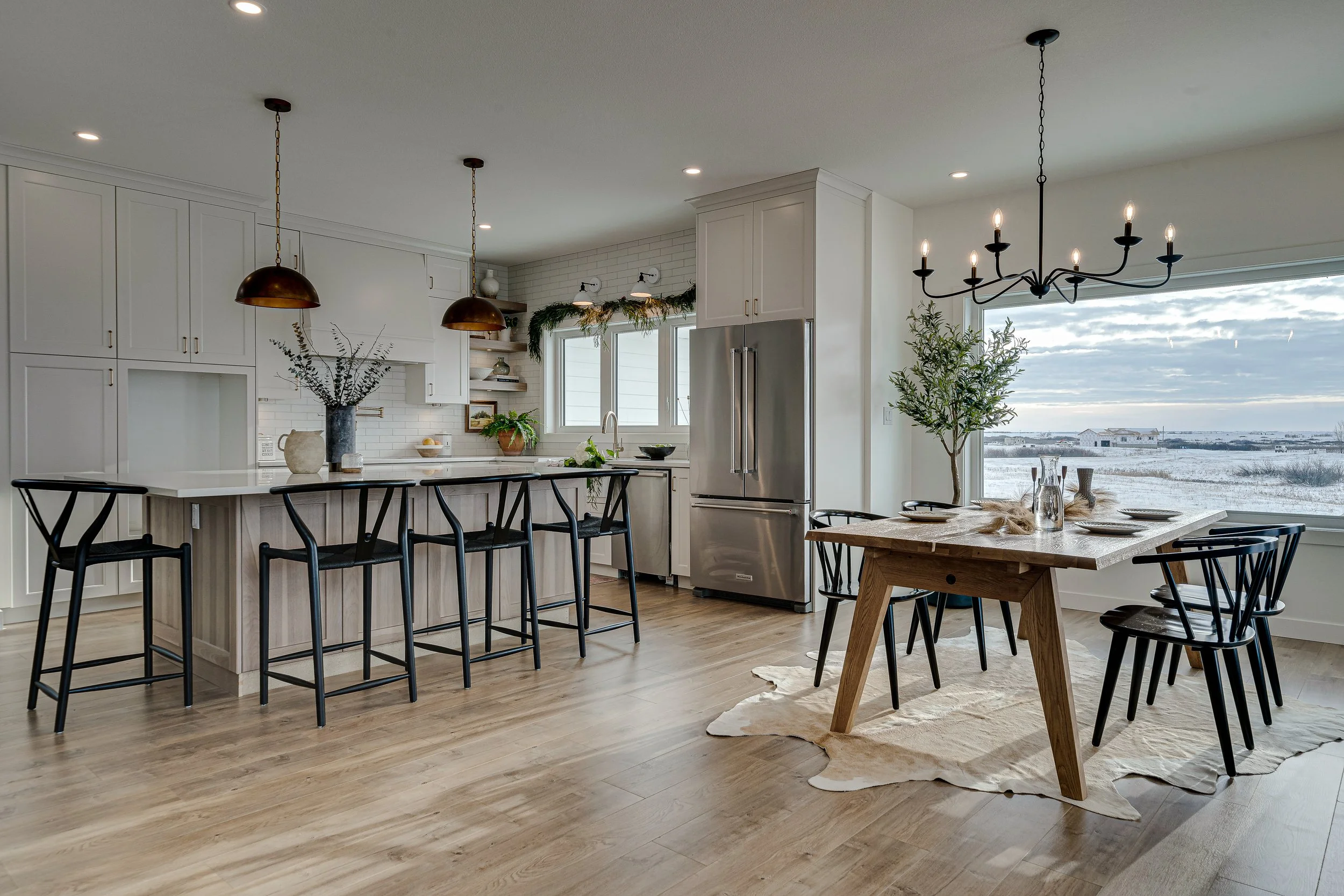 Modern kitchen with white cabinets, stainless steel refrigerator, black bar stools at a kitchen island, and a dining table with black chairs. Large window showcasing a snowy landscape outside. Decor includes vases, plants, and a chandelier.