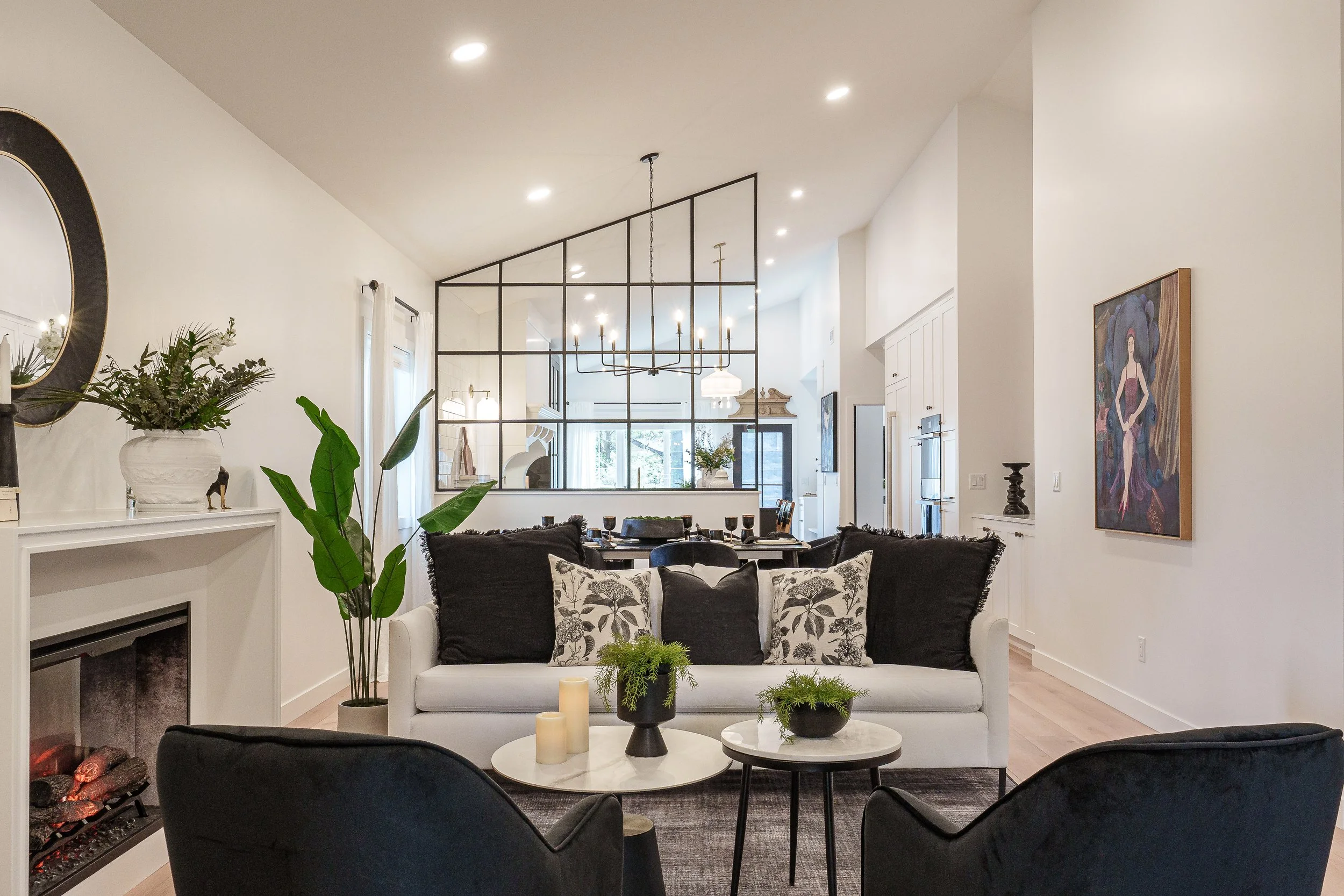 Open-concept living room and dining area with modern decor, white walls, pendant and chandelier lighting, plants, and a framed painting.