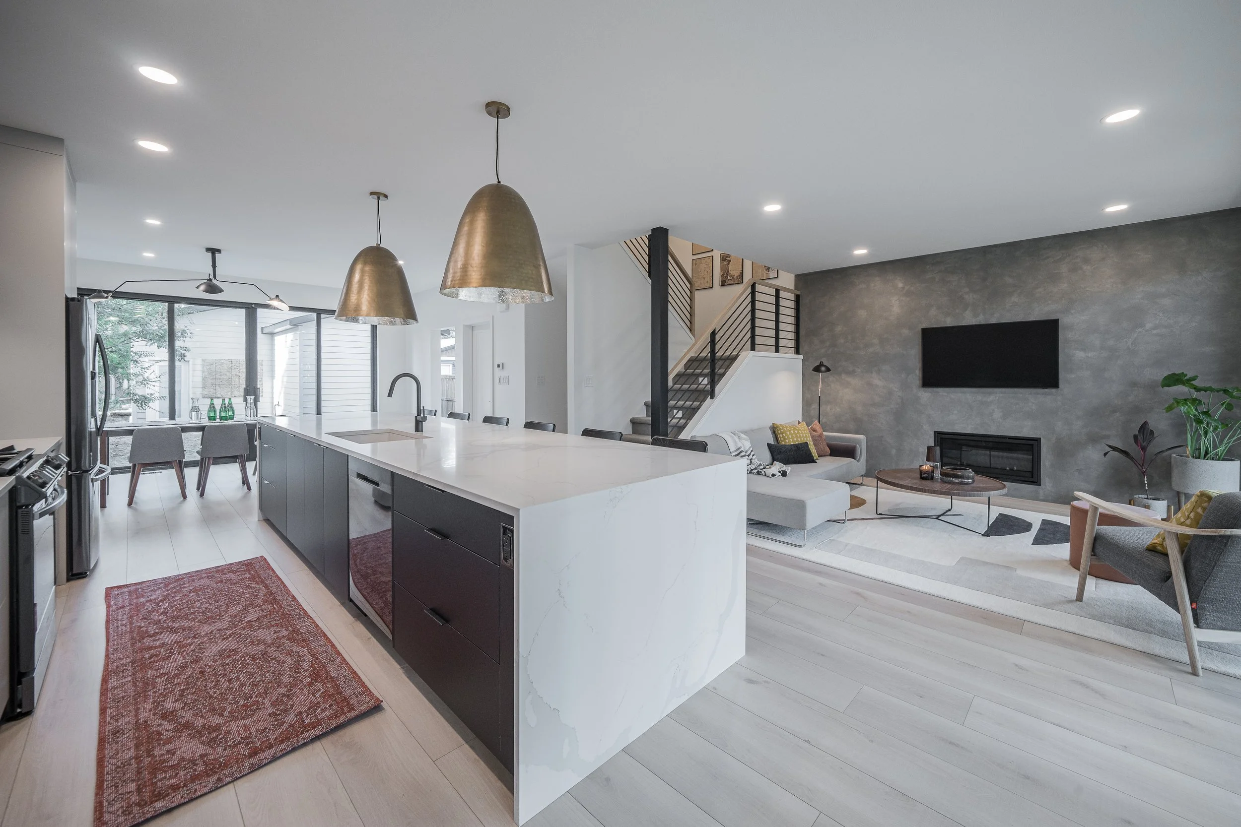Modern open-concept living room and kitchen with white walls, light wood flooring, and recessed lighting. The kitchen features a large island with a waterfall marble countertop and black cabinetry. Two silver pendant lights hang above the island. The living area has a gray textured accent wall, a mounted flat-screen TV, a fireplace, and a gray sofa with colorful pillows. Large windows and sliding glass doors let in natural light.