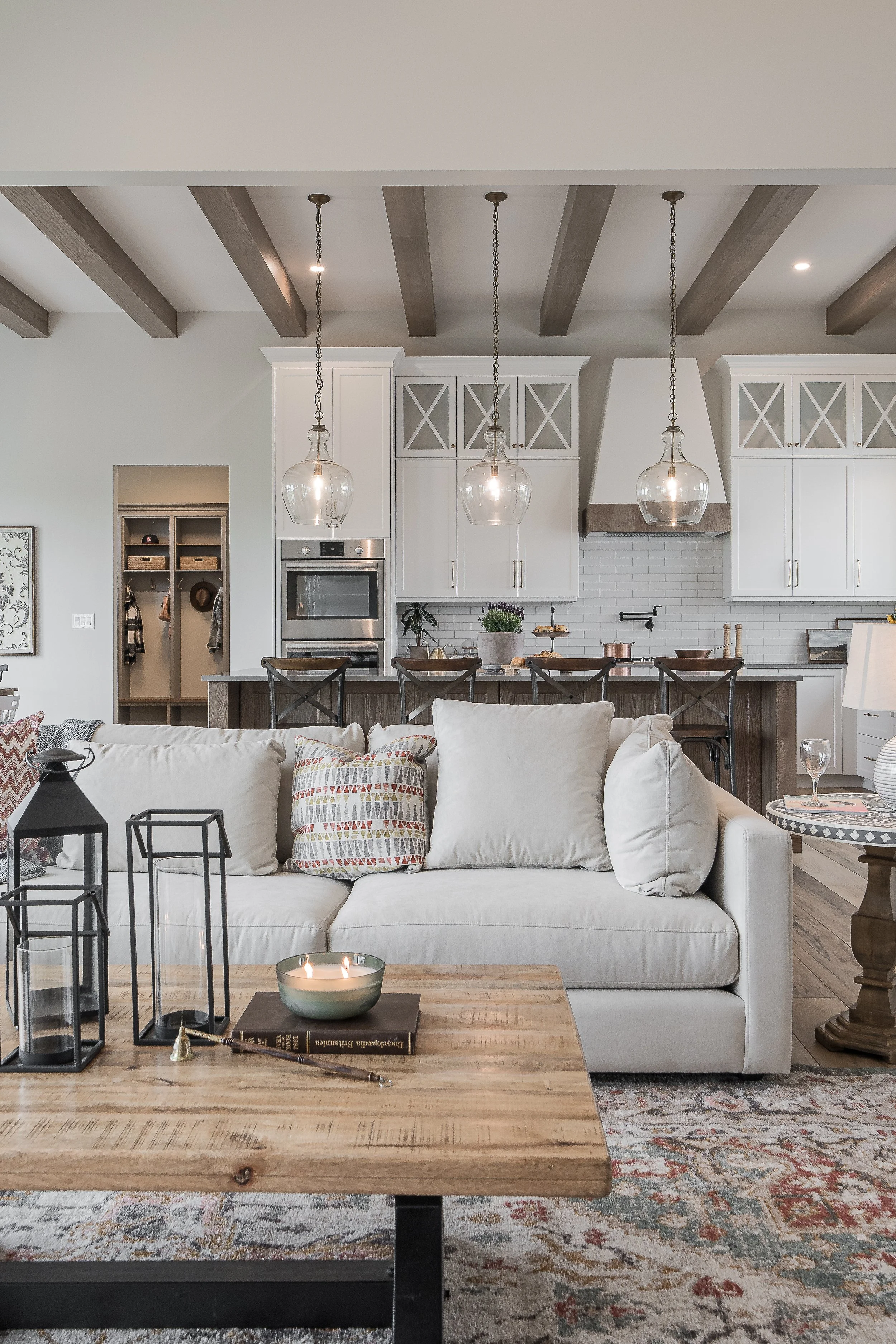 Open-concept kitchen and living room with white cabinets, wood ceiling beams, pendant lights, a kitchen island, and a white sofa with decorative pillows.