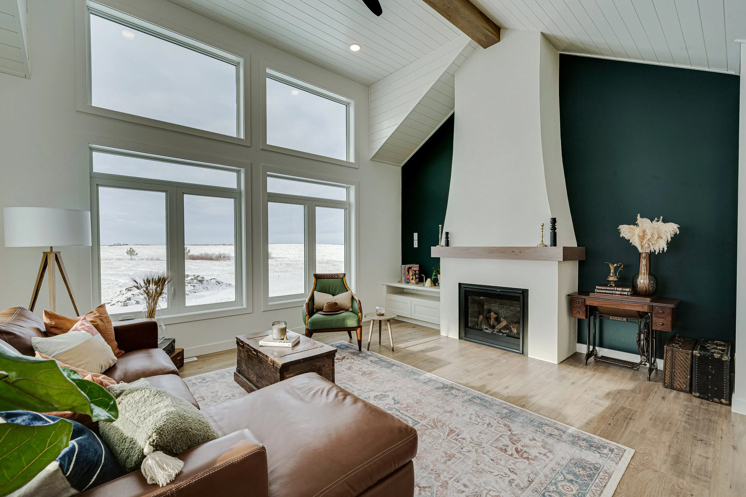 Living room with large windows overlooking snowy landscape, a leather sofa with colorful pillows, a green armchair, a coffee table, a fireplace, and a decorative vase with pampas grass.