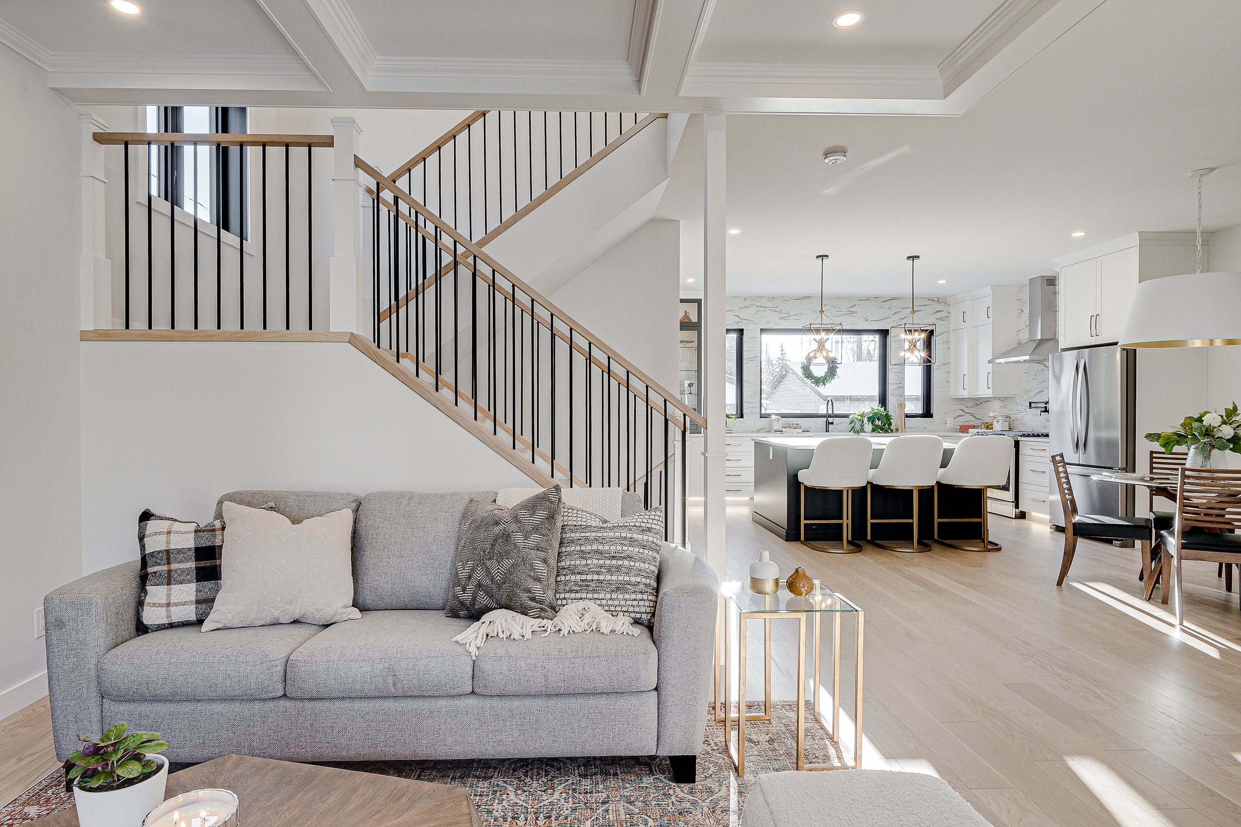 Open-concept living room and kitchen with staircase, white walls, large windows, and modern furniture.
