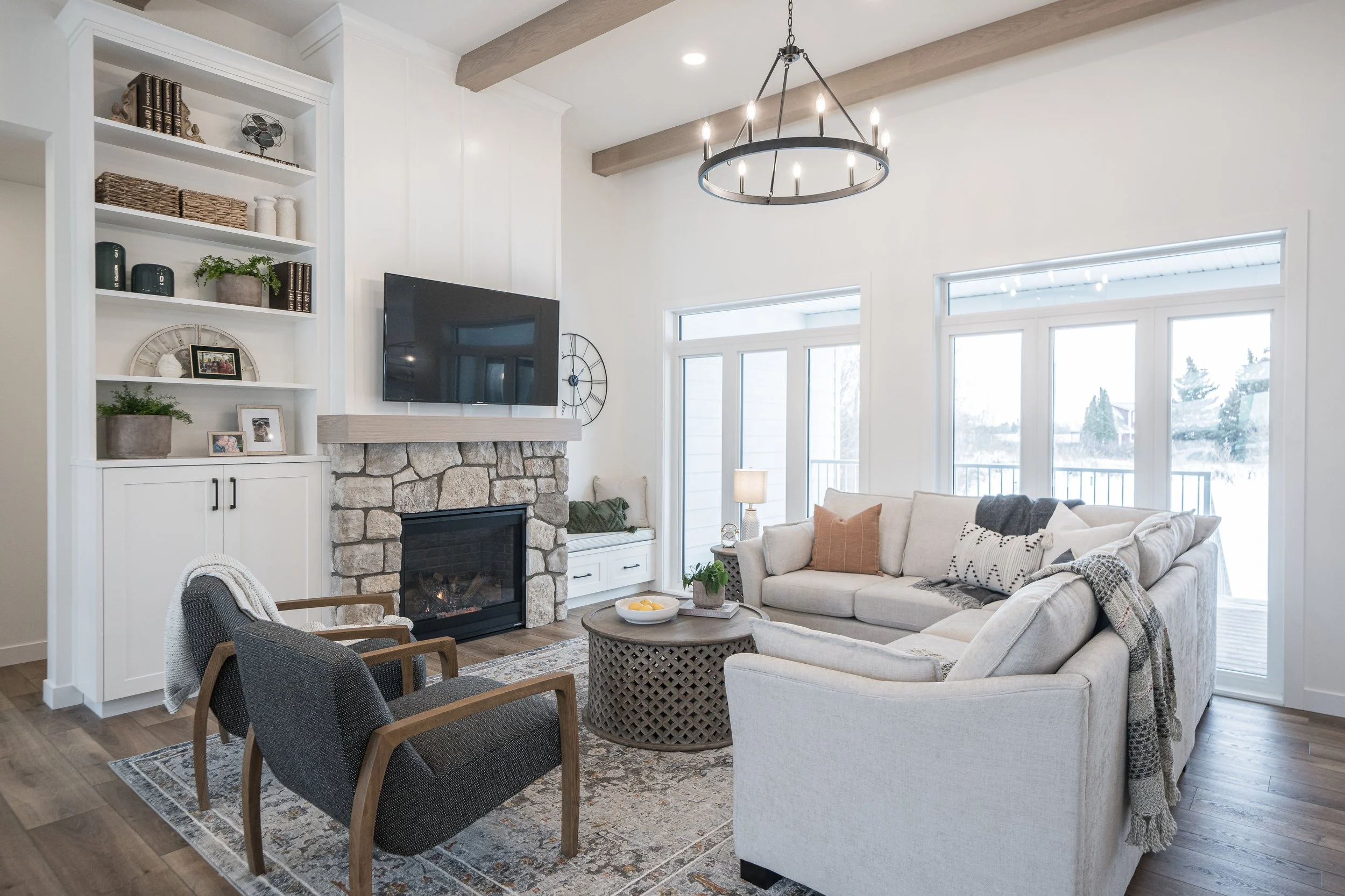 Living room with a stone fireplace, a wall-mounted TV, white built-in shelves, a round chandelier, large windows, a cream-colored sofa with decorative pillows, dark wooden chairs, a round coffee table with a bowl of lemons, and a patterned area rug.
