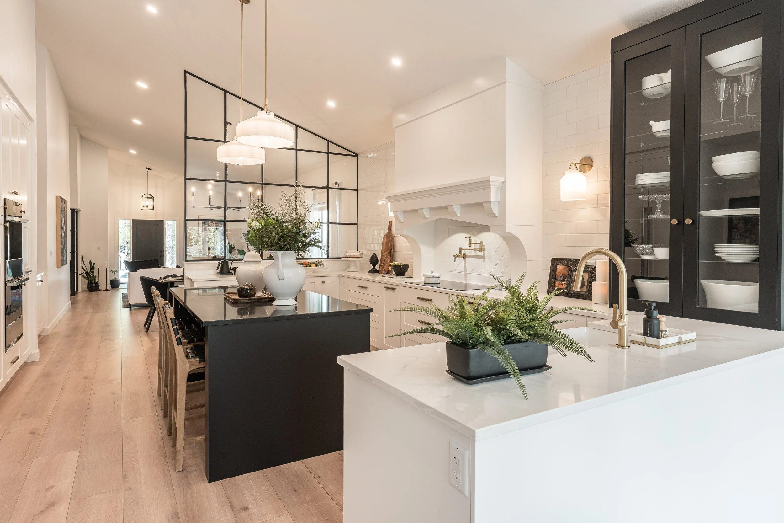 A modern kitchen with white cabinets, a black kitchen island, a large white oven, a glass-paneled black cabinet, hanging light fixtures, a large white vase with flowers on the island, and a white fireplace with gold fixtures.