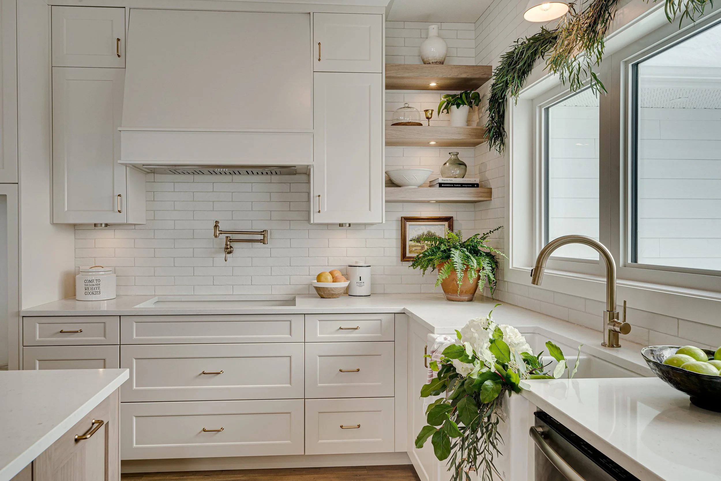 A modern kitchen with white cabinets, open wooden shelves, a white brick backsplash, and a large window. Decor includes plants, a white bowl with fruit, and decorative items.