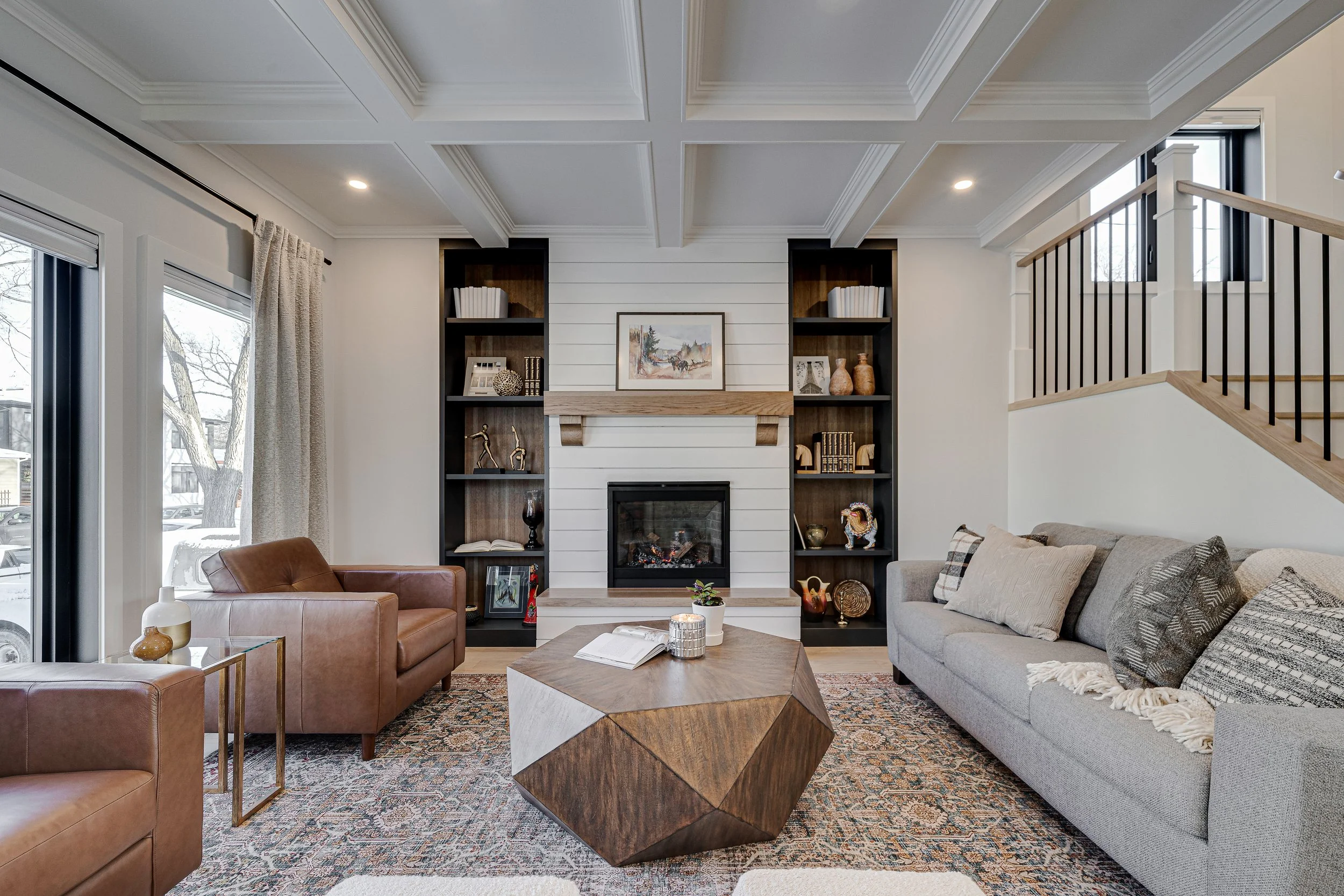 Living room with a fireplace flanked by built-in bookshelves, a gray sofa with pillows, a brown armchair, a geometric wooden coffee table, and large windows with curtains and black frames.