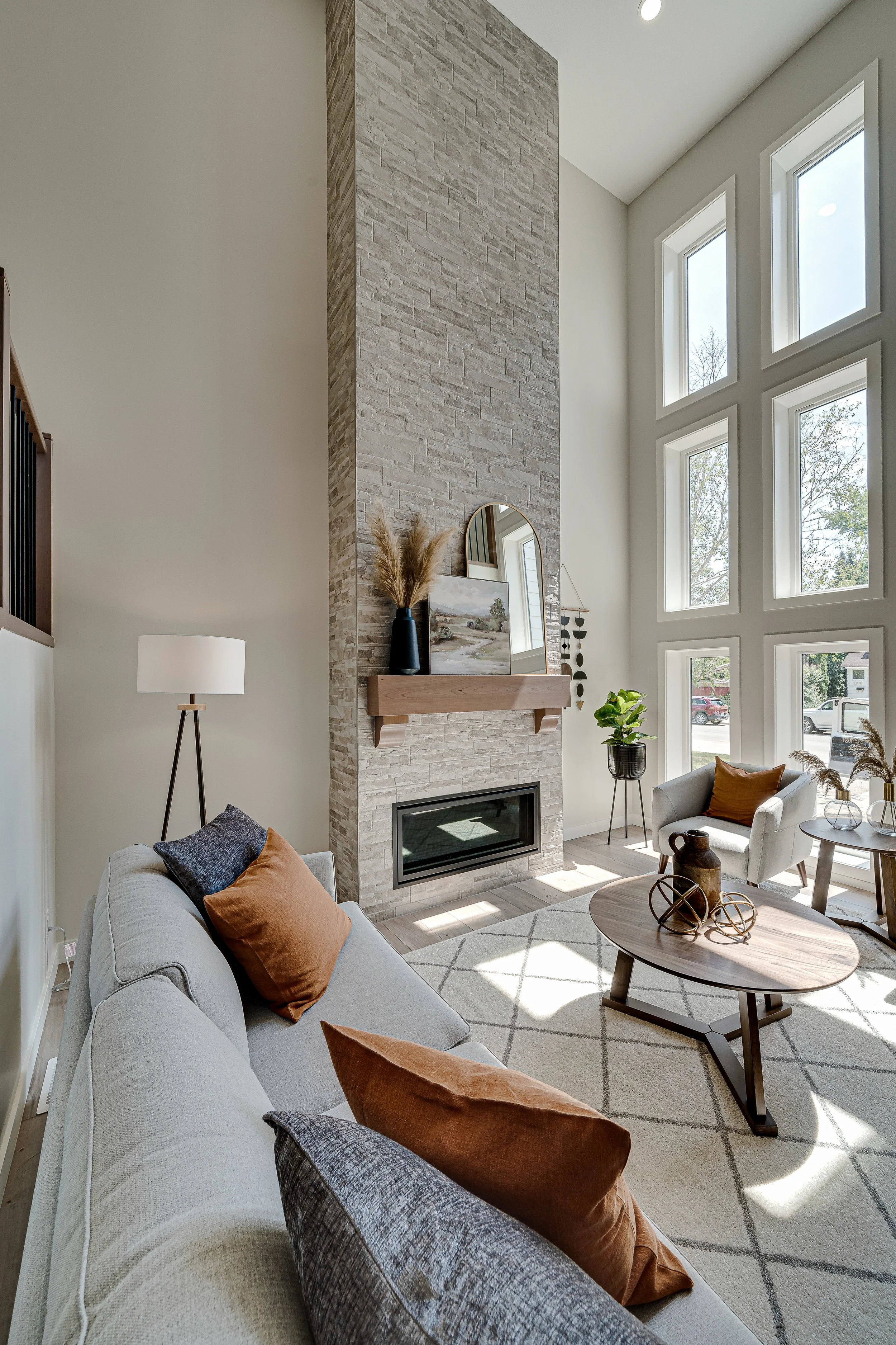 Living room with a beige sofa adorned with rust and dark gray pillows, a wooden coffee table with decorative items, a tall black floor lamp, a fireplace with a beige brick chimney, a small plant, a framed painting, a mirror, and a window wall letting in natural light.