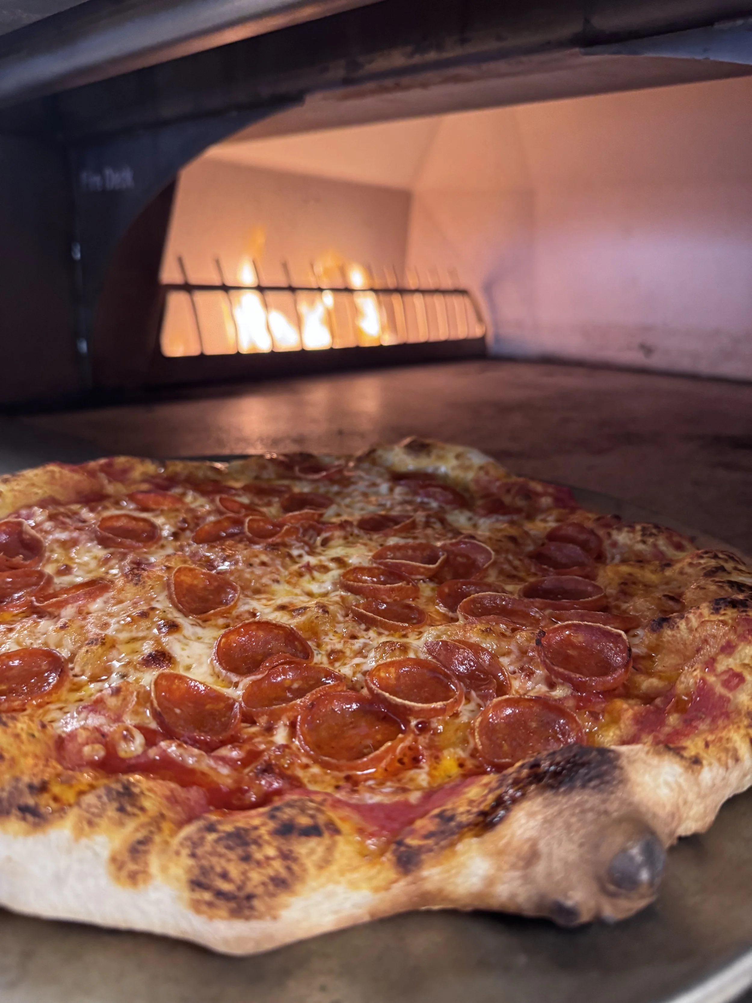 A pepperoni pizza inside a wood-fired oven with flames in the background.