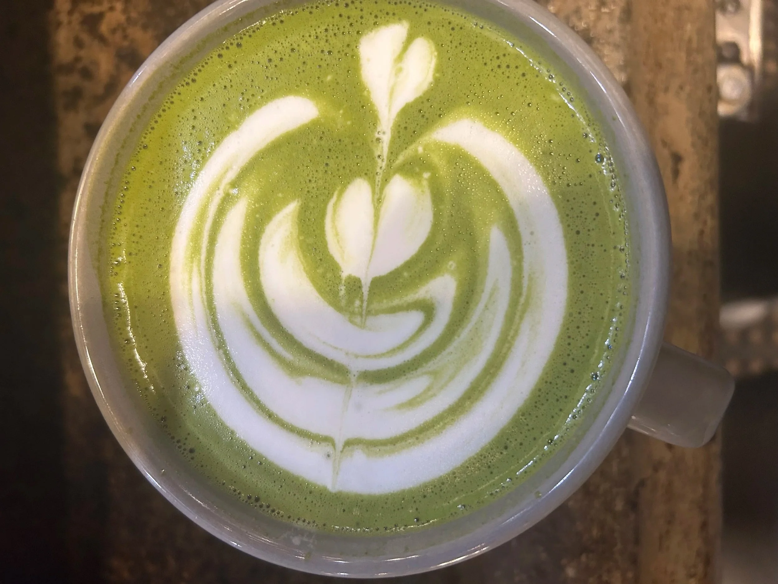 A top view of a latte in a glass cup with green matcha tea, topped with white and green latte art in a floral pattern. Vintage Coffeehouse