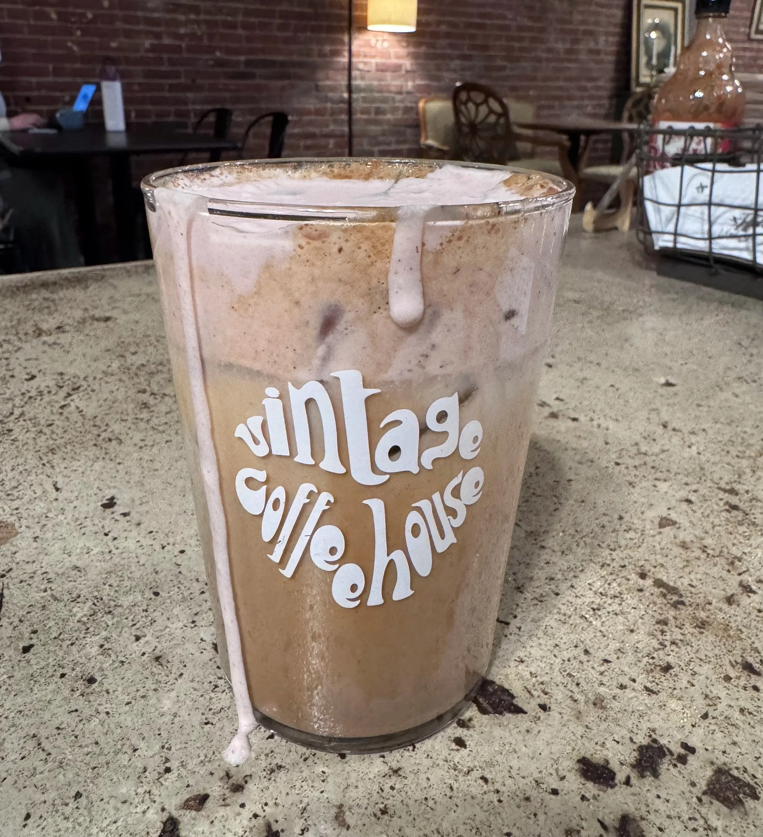 A tall glass with the words 'vintage coffee house' printed on it, filled with iced coffee topped with foam and some drips on the outside, placed on a speckled countertop in a cozy cafe setting. Vintage Coffeehouse
