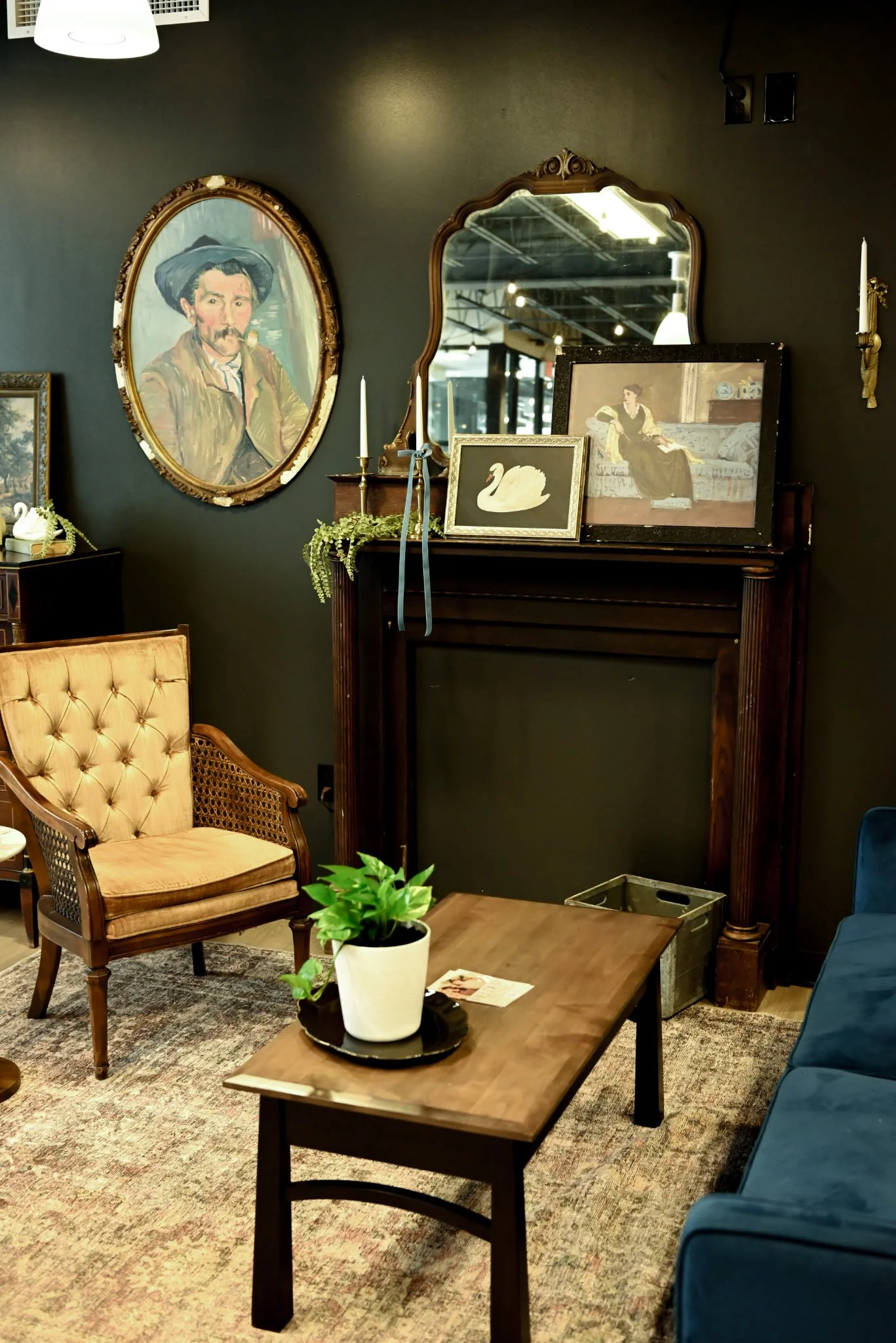 Interior of a vintage-style living room with framed artwork, a wooden fireplace mantel, a side table, a chair, a plant on a coffee table, and a dark wall.
