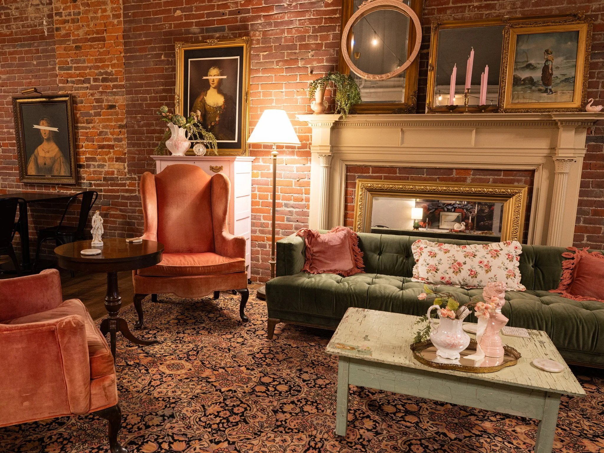 A cozy living room with exposed brick wall, vintage furniture including a green velvet sofa with floral and pink pillows, a high-backed pink armchair, a dark wooden round side table, and a distressed white coffee table decorated with pink ceramic vases. The brick wall features framed artwork, and a white mantel with paintings, candles, and a round mirror. A floor lamp provides warm lighting, and the room has a patterned rug.
