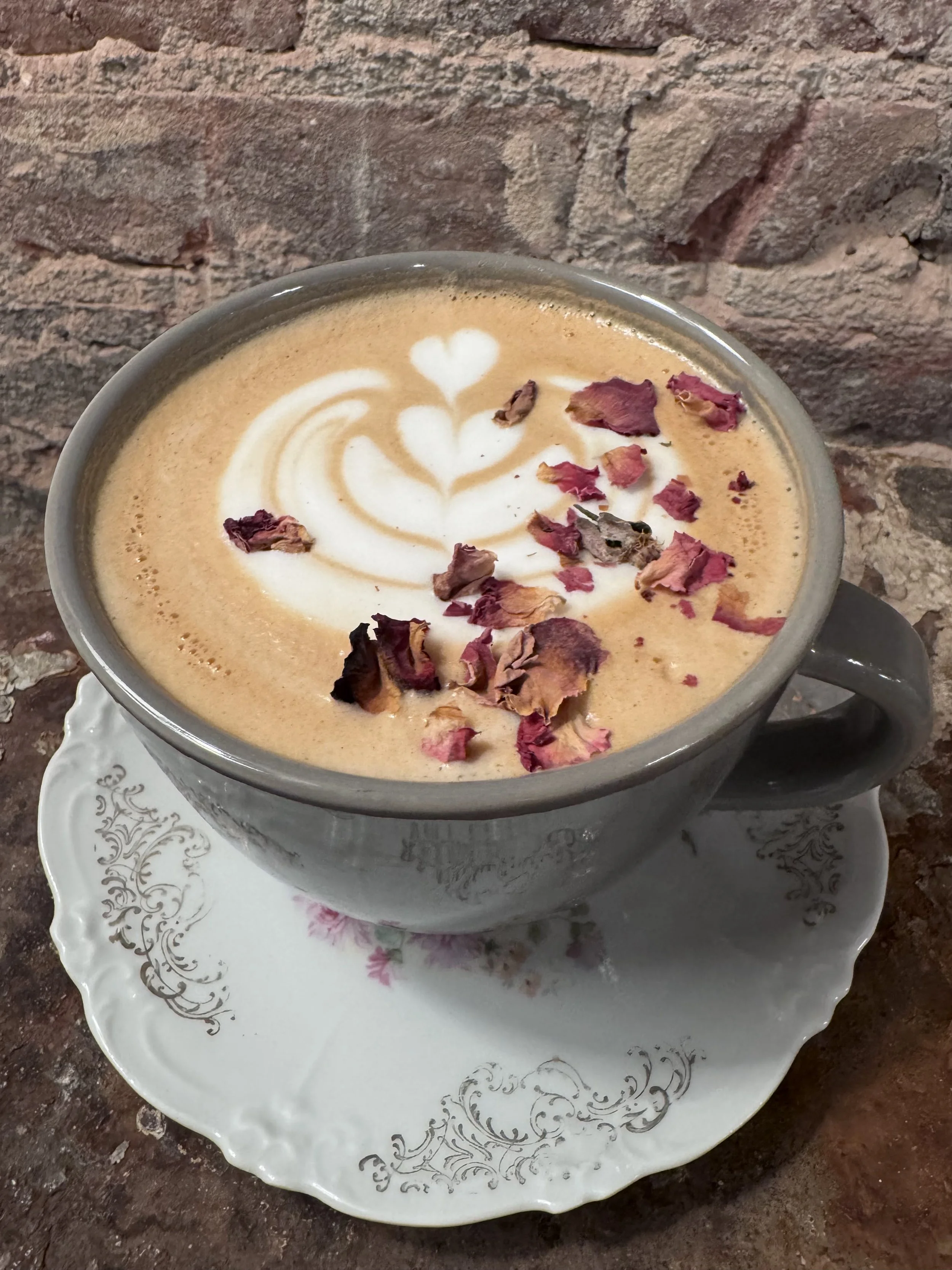 A cup of coffee with latte art and dried rose petals on top, placed on a decorative white saucer on a brick wall background. Vintage Coffeehouse