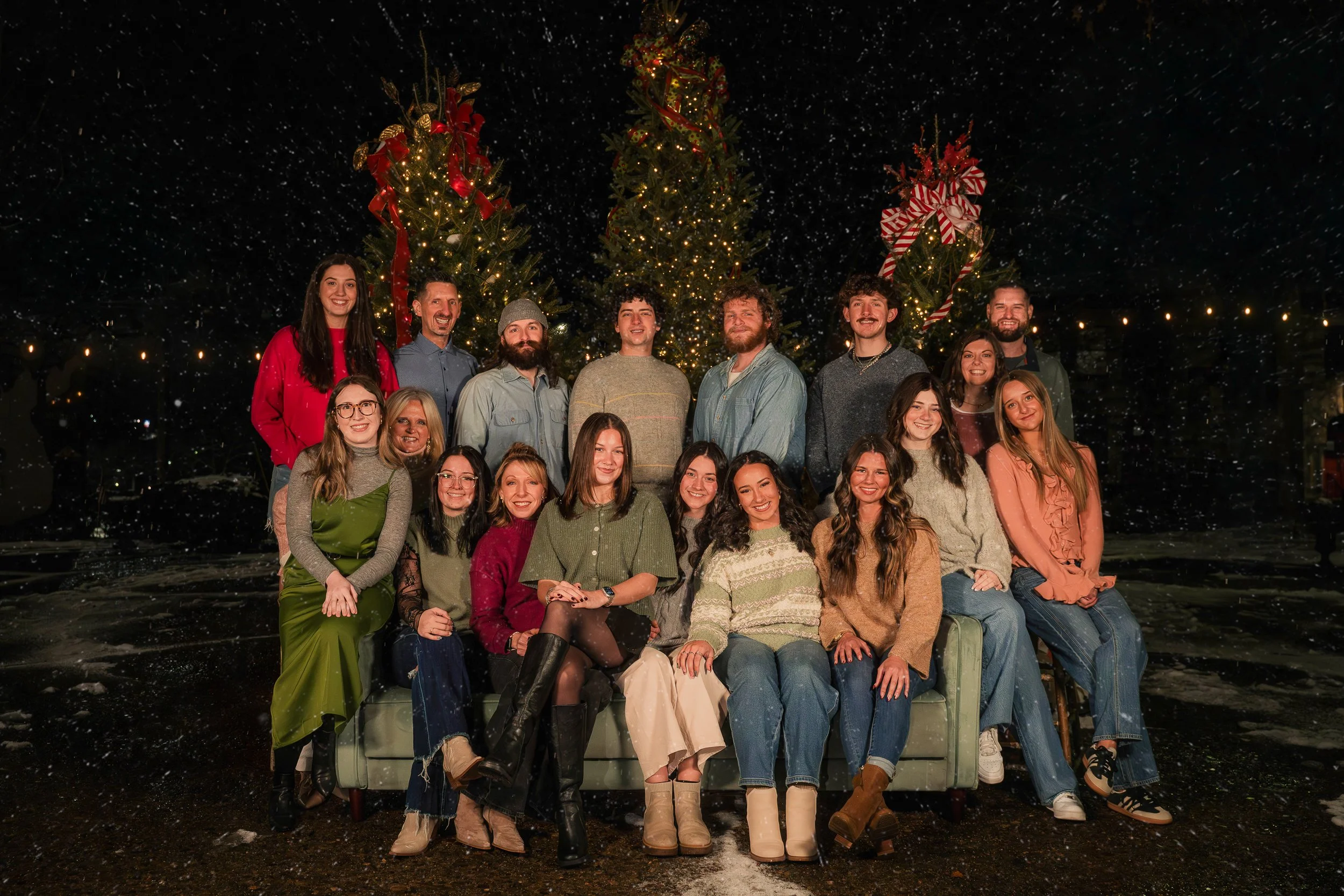 A group of 17 people celebrating Christmas outdoors at night, with decorated Christmas trees and falling snow.