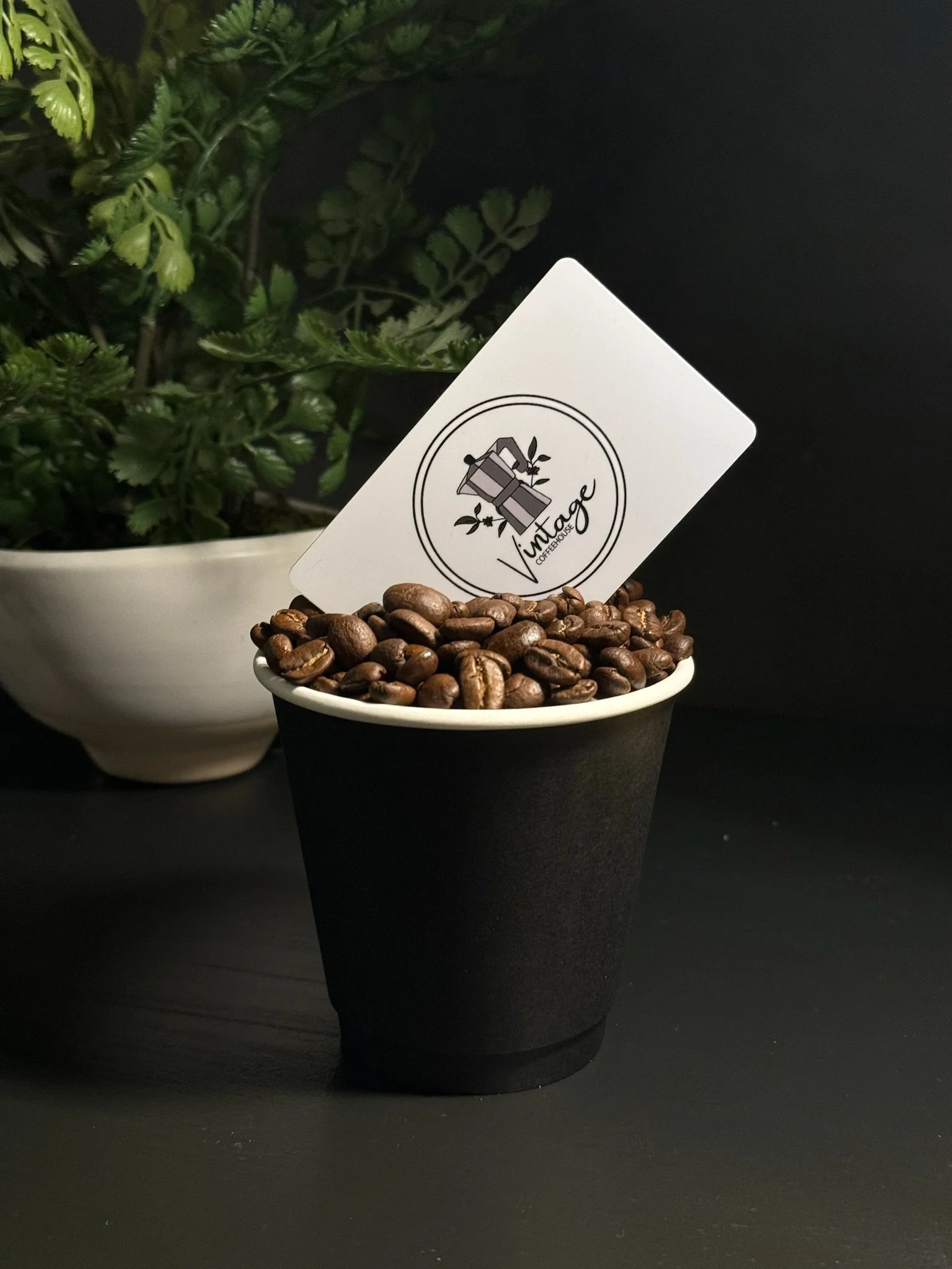 A black cup filled with roasted coffee beans and a card with a logo that reads 'Vintage' from a coffee house, with green plants in the background.