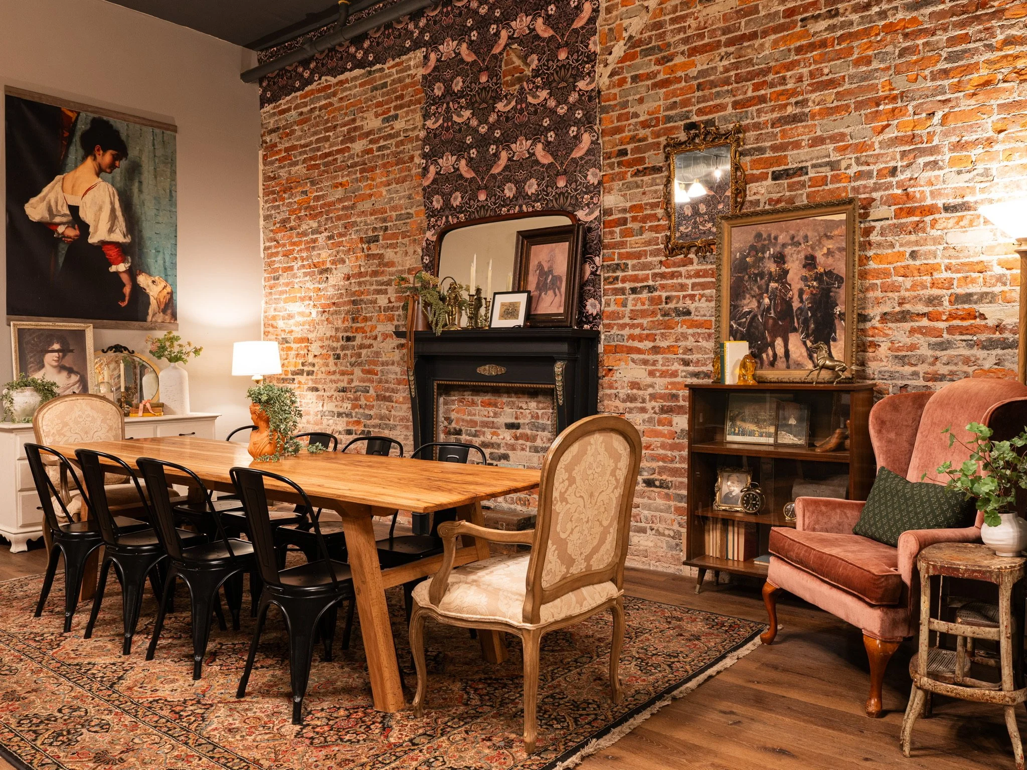 A cozy dining room with a large wooden table surrounded by black chairs and an upholstered armchair. The room features an exposed brick wall decorated with paintings, a mirror, and vintage-style furniture, creating an eclectic and warm atmosphere.