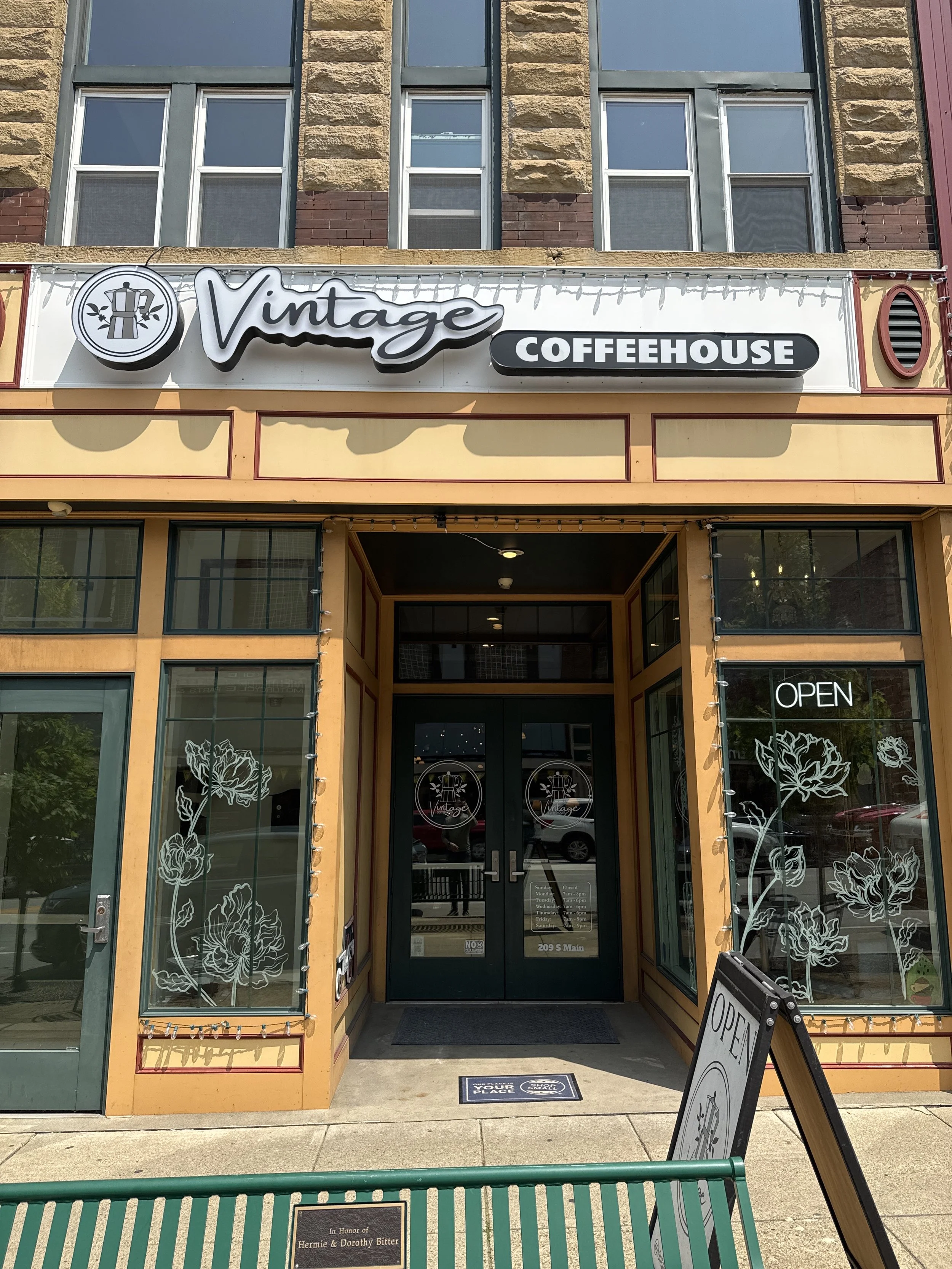 The exterior of a coffee shop called Vintage Coffeehouse with yellow and brown trim, large glass windows decorated with white floral designs, and an 'OPEN' sign on the window. A sidewalk sign displays the word 'OPEN'.