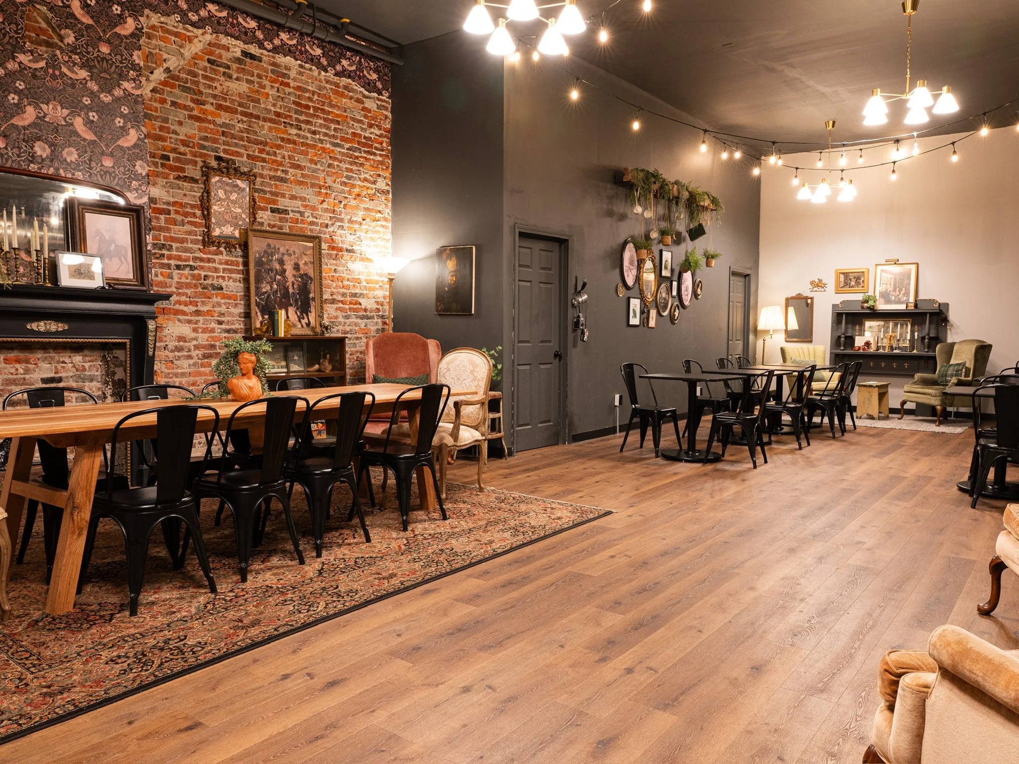 Spacious lounge or event space with wooden floors, brick and gray walls decorated with vintage pictures and plants. There are armchairs, tables, and chairs, with string lights hanging from the ceiling creating a cozy atmosphere.