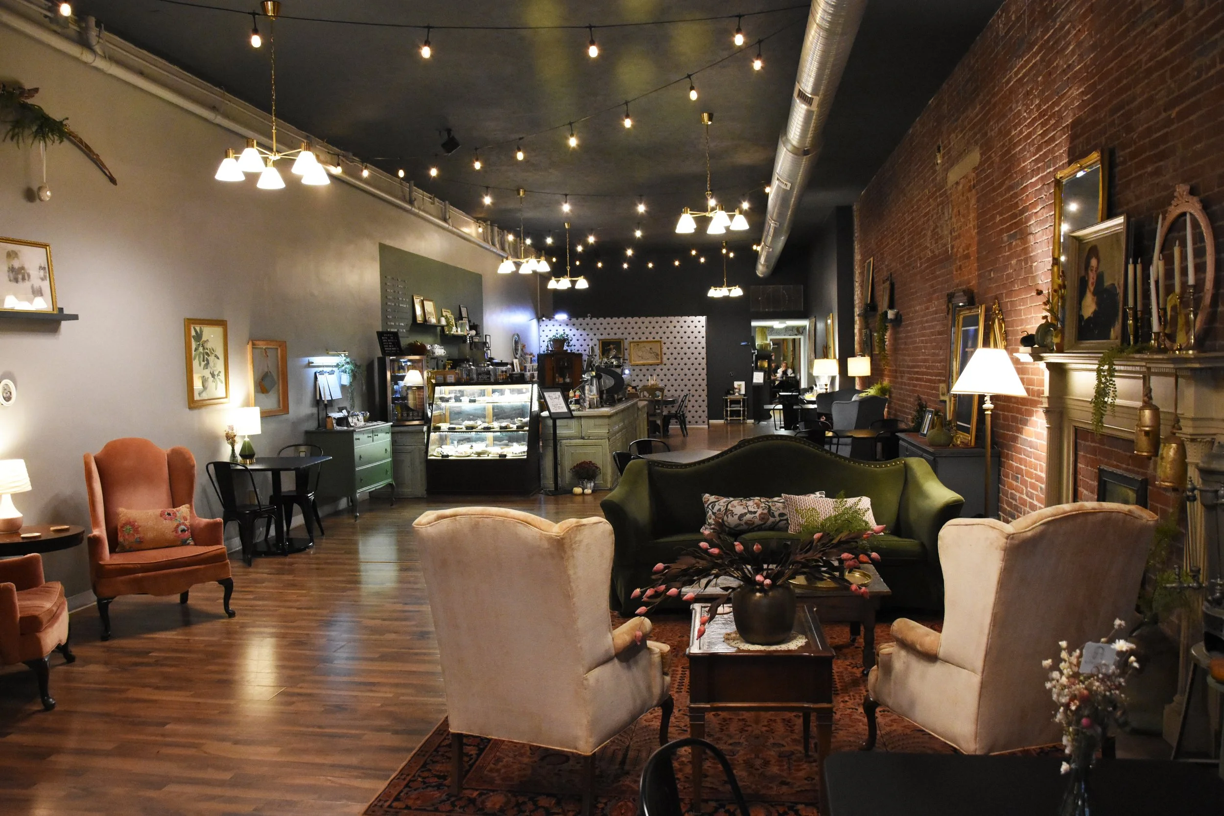 Interior of a cozy cafe or lounge with vintage decor, including velvet armchairs, framed pictures, decorative lamps, and a brick wall, with a coffee display case and ambient string lights overhead.