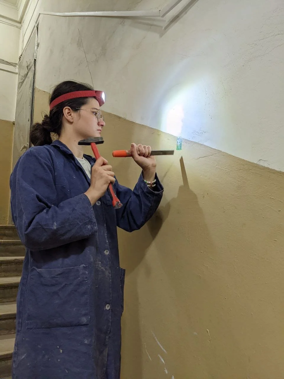 A woman wearing a blue work coat, glasses, and a red headlamp, using a hammer and chisel to remove paint or plaster from a wall.