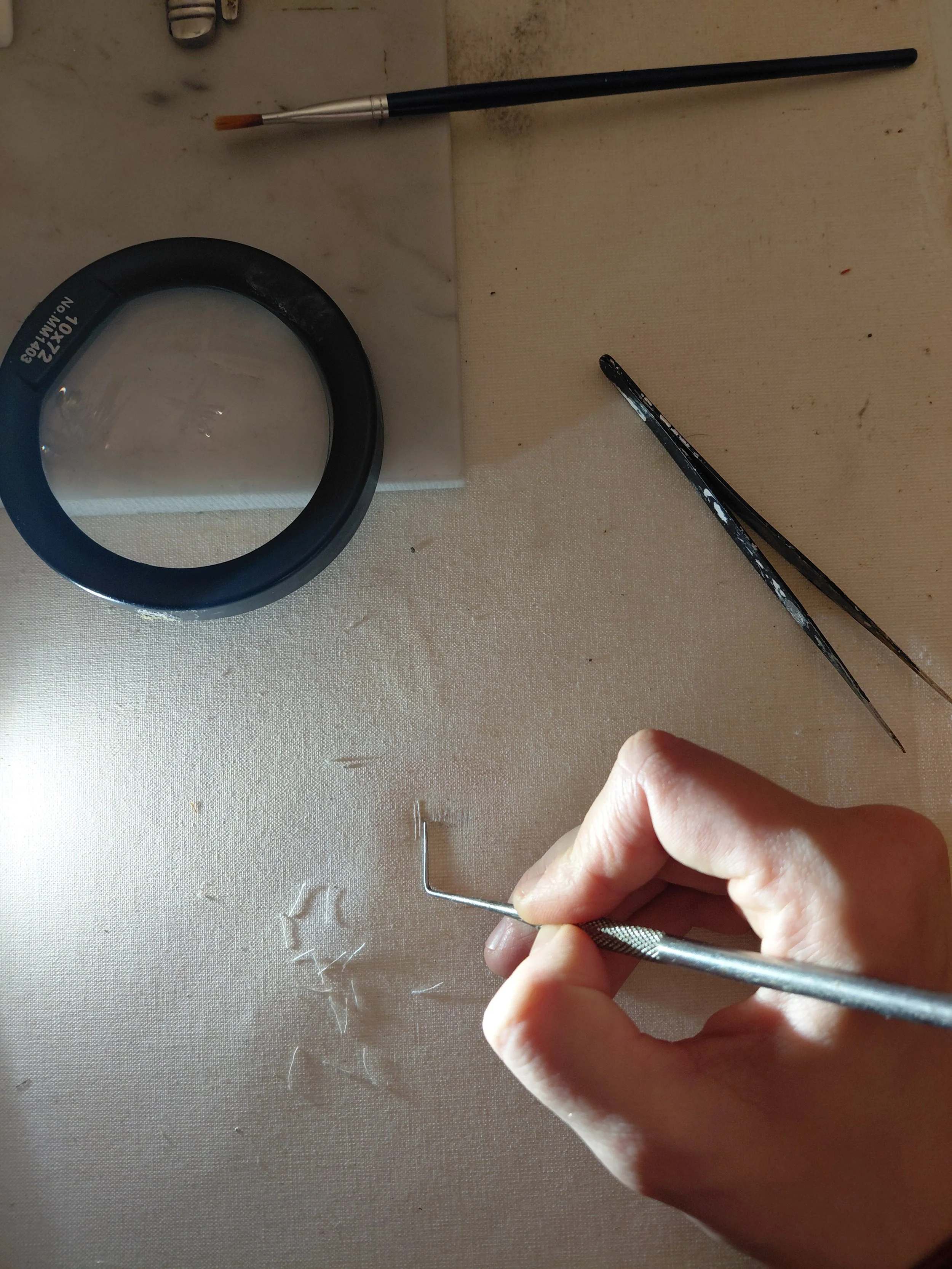 A hand holding a small metal hook tool, working on a canvas surface with torn brush bristles scattered on it. Various art tools are visible, including a magnifying glass, tweezers, a paintbrush, and a stylus with a black handle.