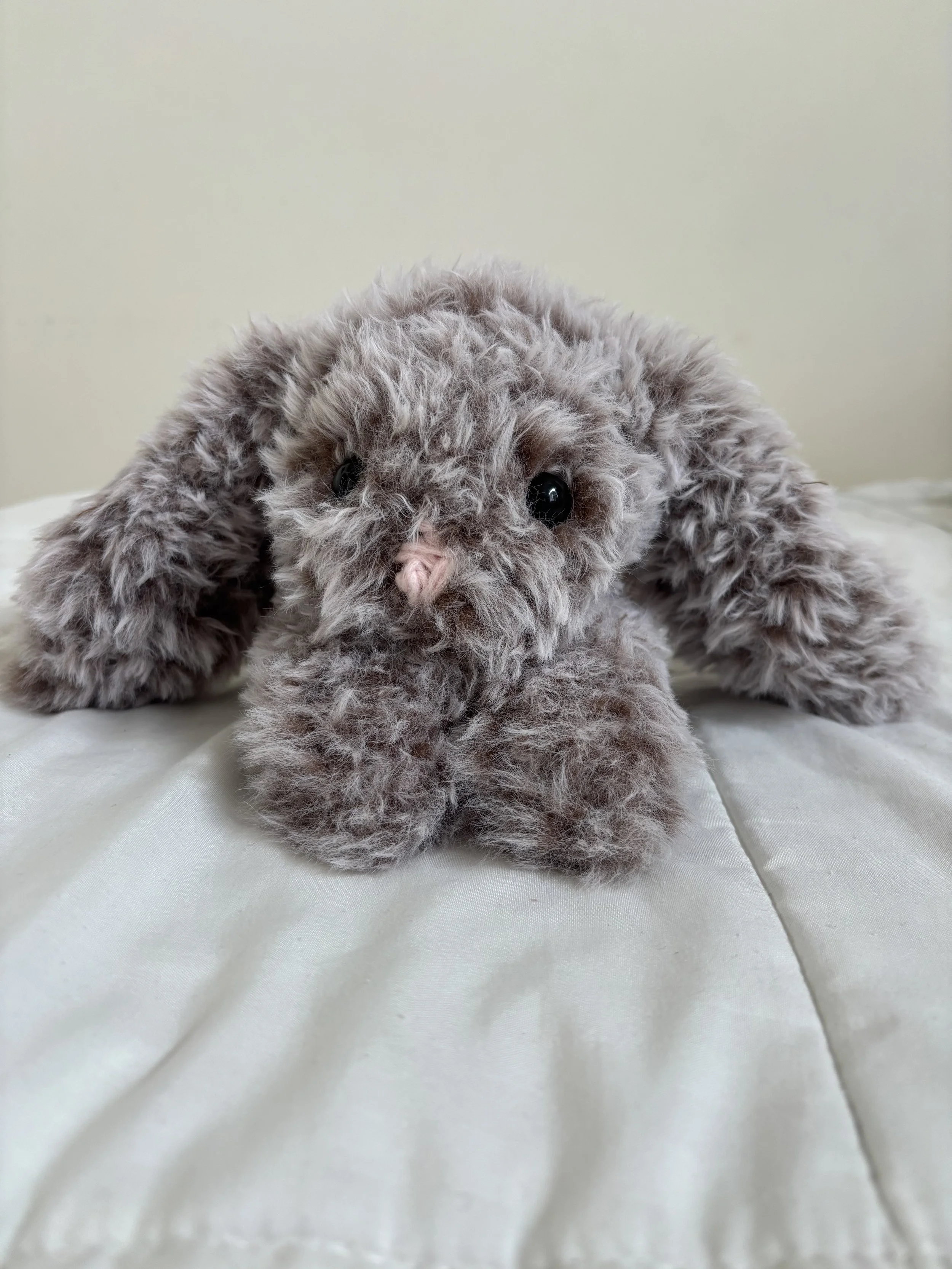 A close-up of a fluffy gray stuffed bunny with floppy ears, shiny black eyes, and a small pink nose, lying on a white fabric surface.