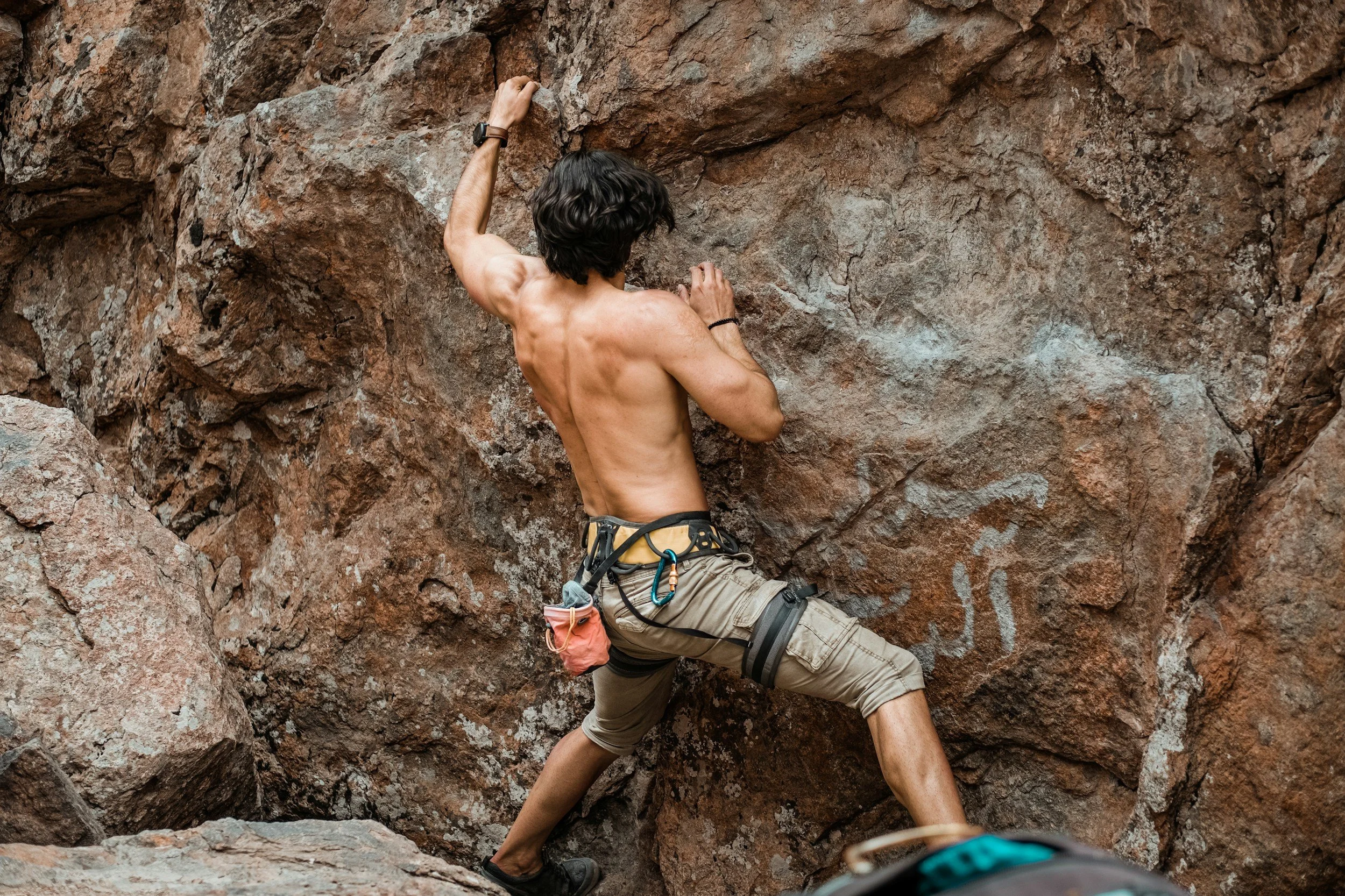 A shirtless male rock climber in beige shorts is ascending a rugged, reddish-brown rock wall, with one hand gripping a hold and the other reaching upward, wearing a climbing harness with carabiners.