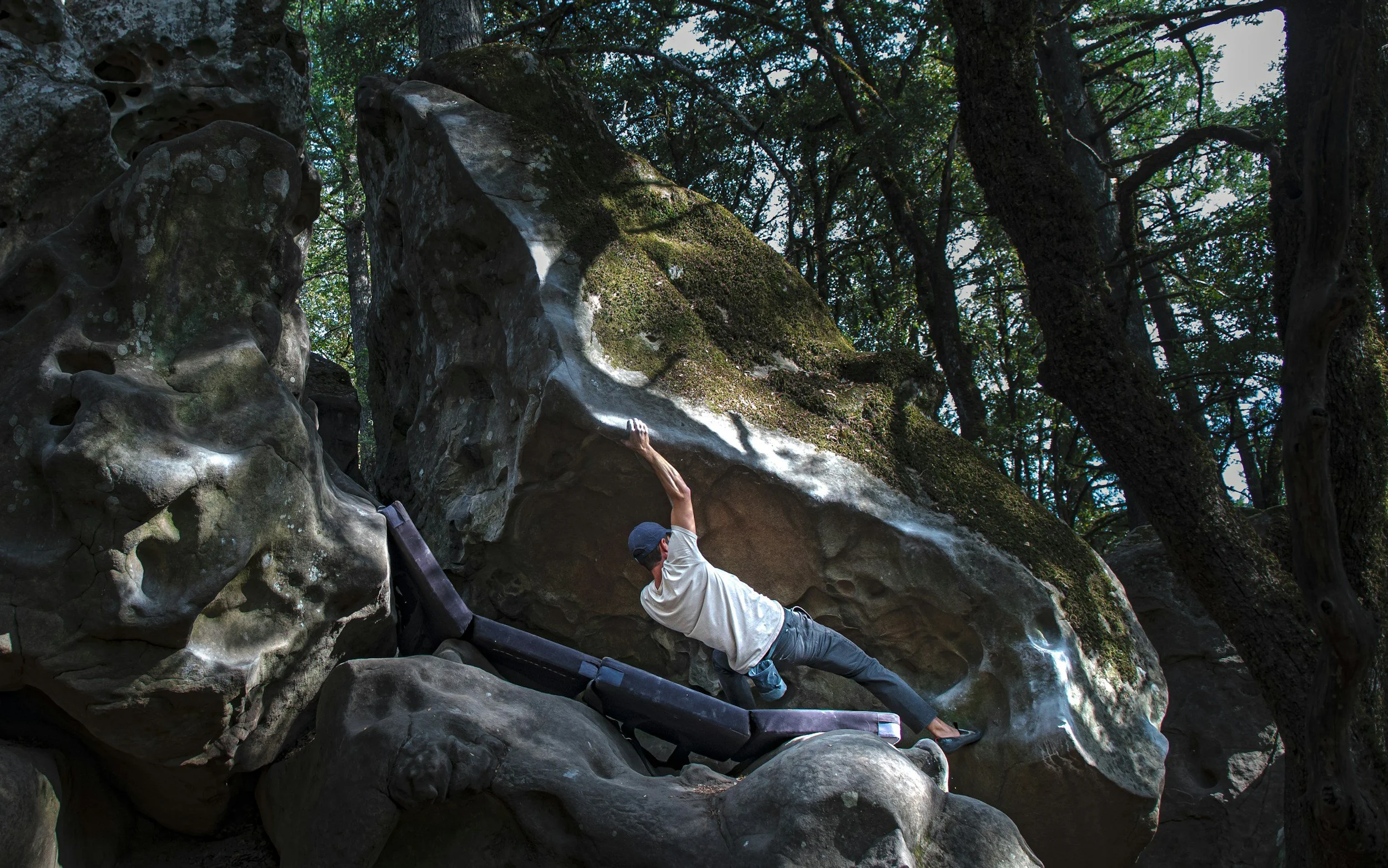 A person wearing a white shirt, black pants, and a hat is bouldering on a large rock in a forest, surrounded by trees and moss-covered rocks.