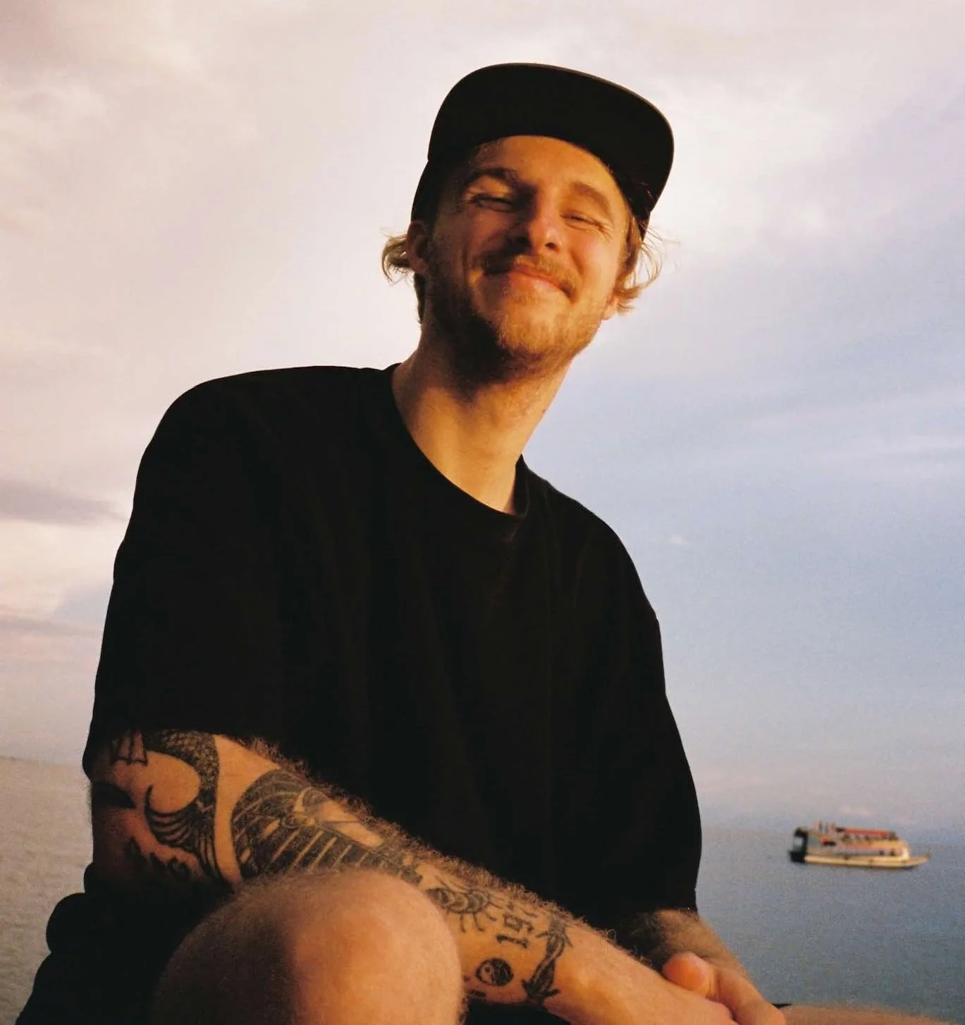 A smiling man with a beard and tattoos on his arm, wearing a black shirt and black cap, sitting outdoors near water with a boat in the background during sunset or sunrise.