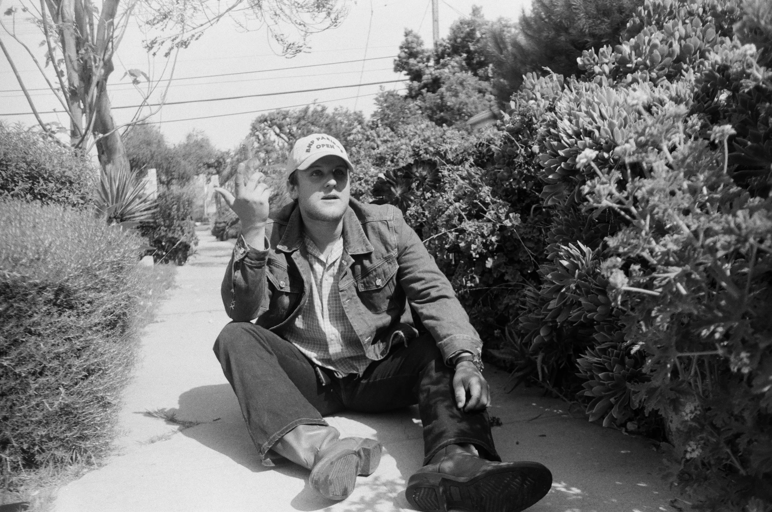 A black and white photograph of a man sitting on a sidewalk, surrounded by bushes and trees, with power lines overhead. The man is wearing a denim jacket, plaid shirt, jeans, boots, and a cap that says 'Mr. Palm Open'. He appears to be looking ahead with a slightly surprised or attentive expression, with one hand raised and the other resting on the ground.