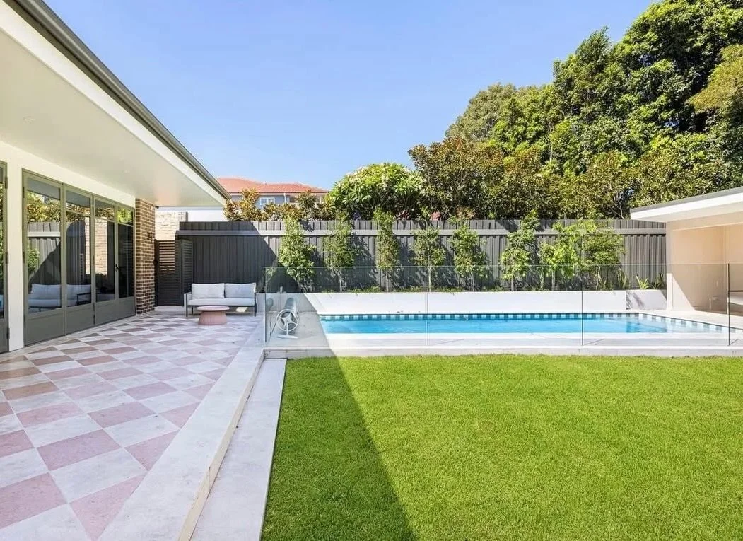 Backyard with swimming pool, patio area with outdoor furniture, green lawn, and fenced garden with trees and shrubs under a clear blue sky.