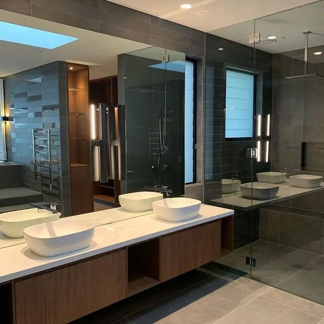 Modern bathroom with double vessel sinks on a wooden vanity, large mirror, walk-in shower with glass enclosure, dark tiled walls, and natural light from a skylight.
