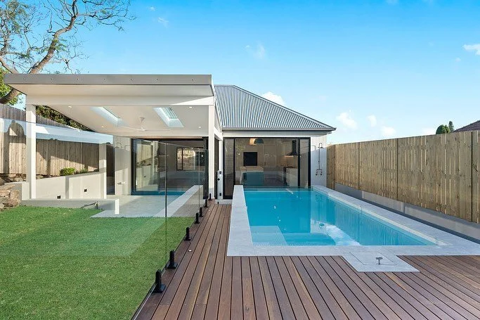 Modern backyard with a rectangular swimming pool, wooden deck, glass fencing, and a small house or pool house with a metal roof.