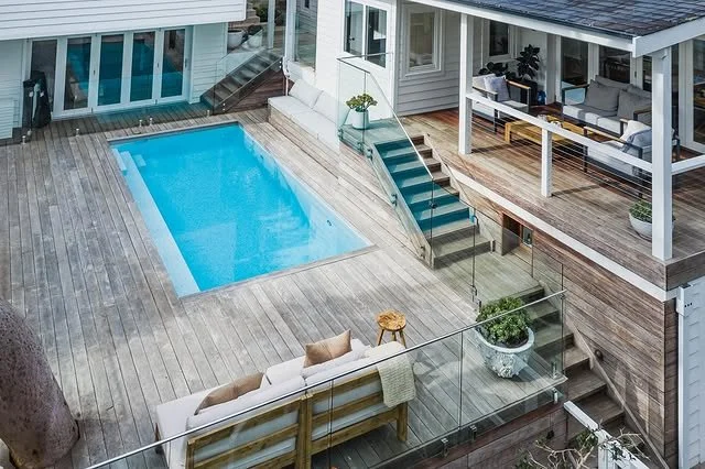 A backyard with a swimming pool, wooden deck, outdoor furniture, and stairs leading to an upper porch with seating area.