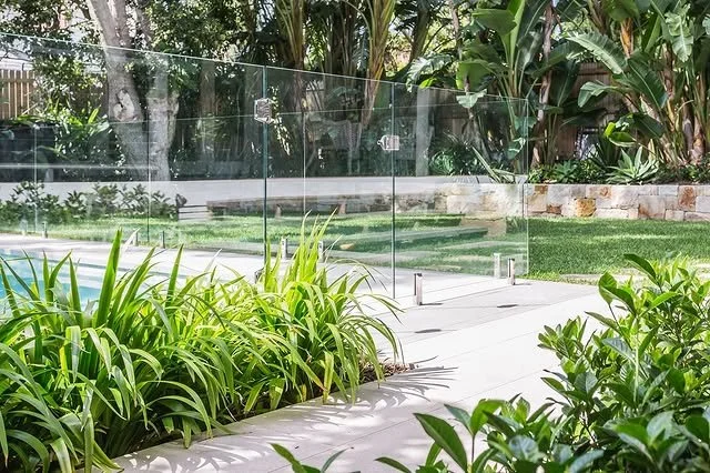 Glass pool fence next to lush green plants and backyard landscaping.