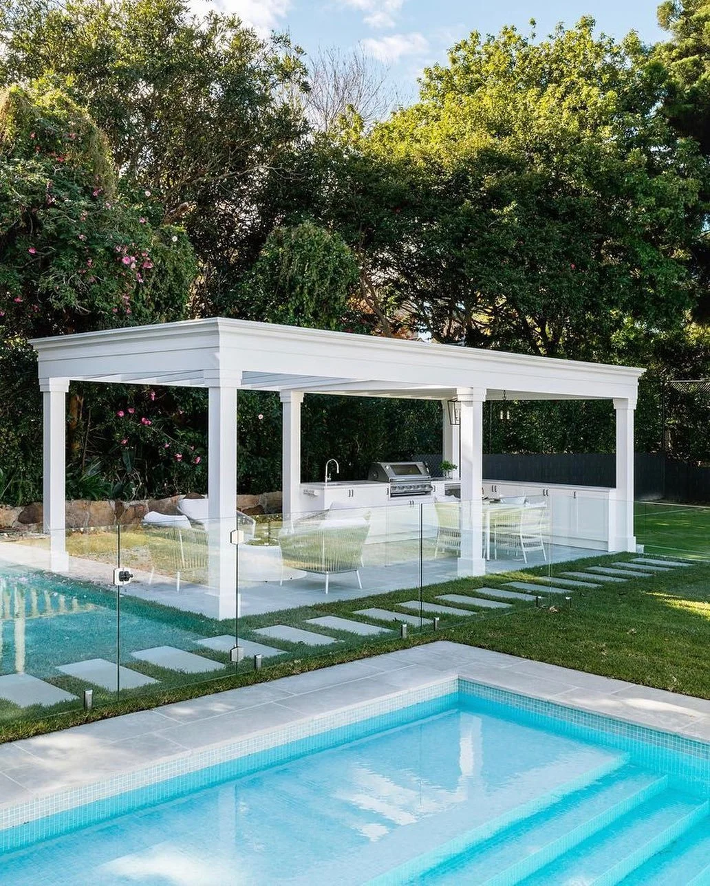 Backyard with in-ground pool and a white outdoor kitchen and seating area under a pergola, surrounded by green trees and plants.
