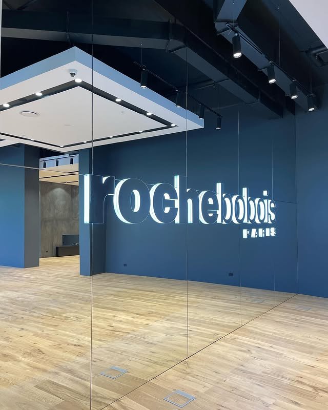 Interior of a modern fitness studio or dance room with a large illuminated 'tRocheobois PARIS' sign on a blue wall, wooden floor, and ceiling lighting.